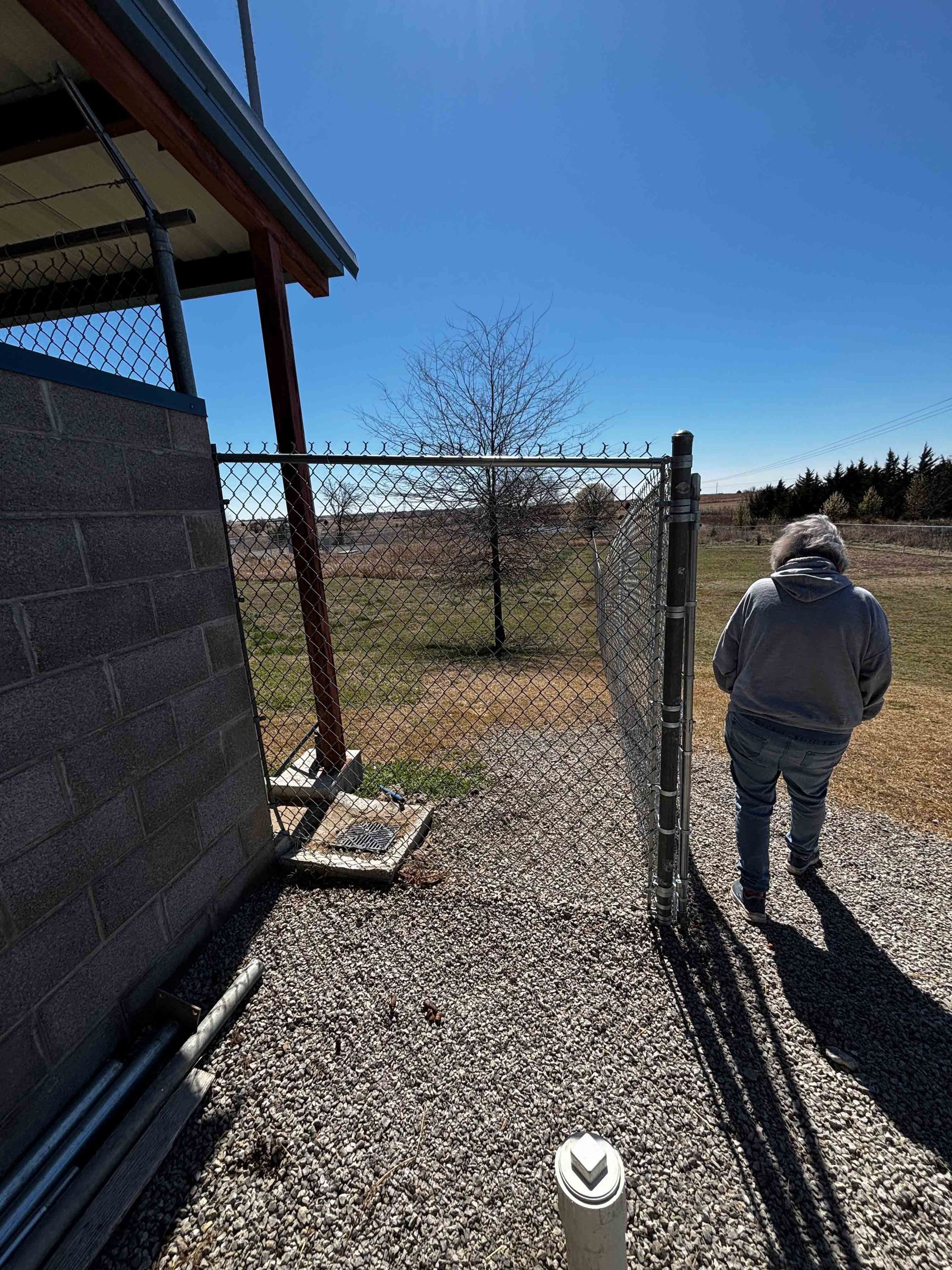 Person in a gray jacket walking by a chain-link fence outside a building on a sunny day