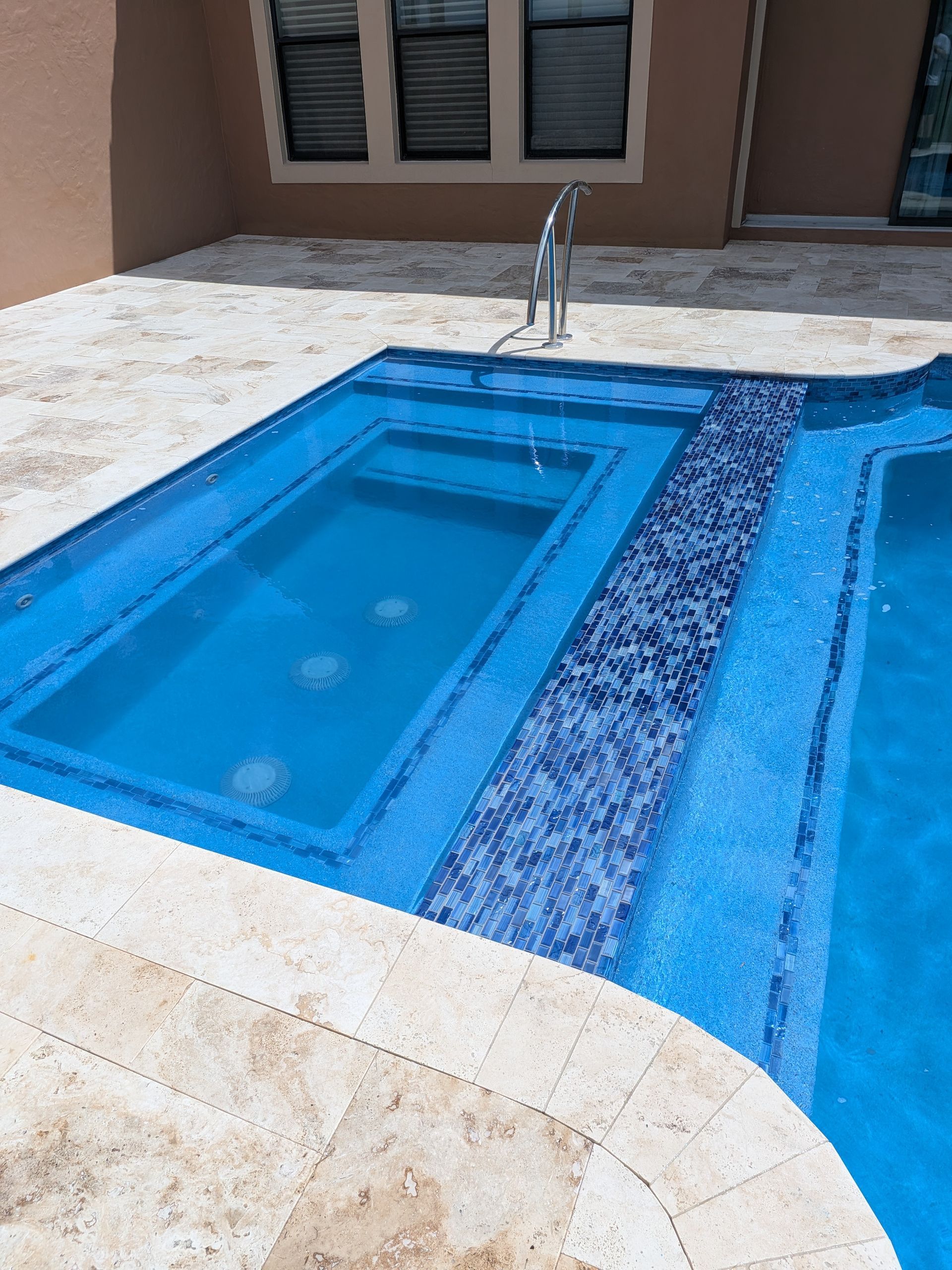 Small blue swimming pool with a spa fountain and tan patio tiles outside a house