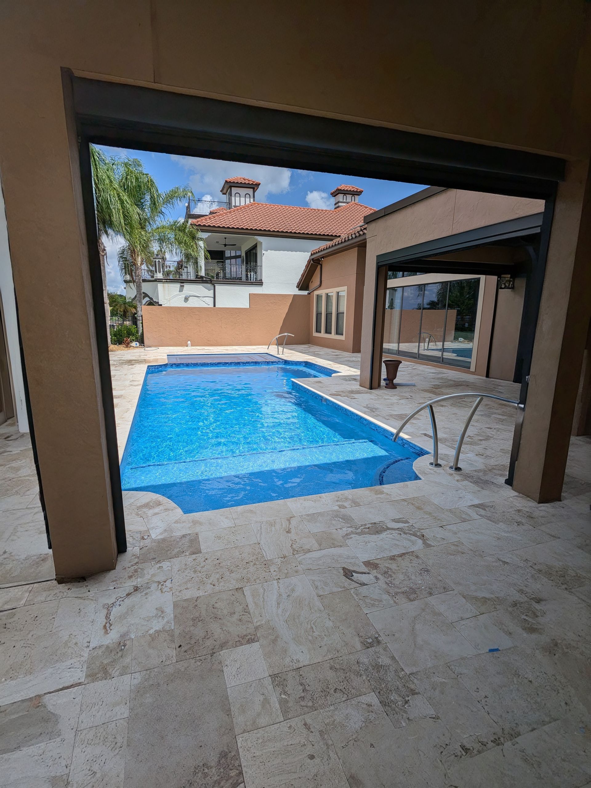Covered patio overlooking a blue swimming pool and stone courtyard, with a house and trees beyond