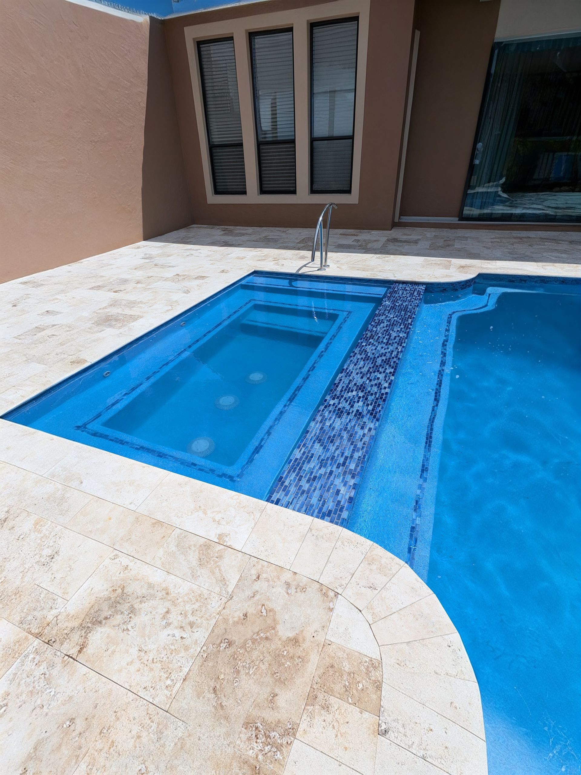 Blue-tiled pool and shallow spa beside a beige patio and sliding glass doors.
