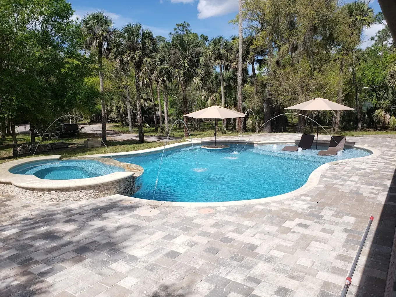 Outdoor resort pool with blue water, two beige umbrellas, and lounge chairs surrounded by trees.
