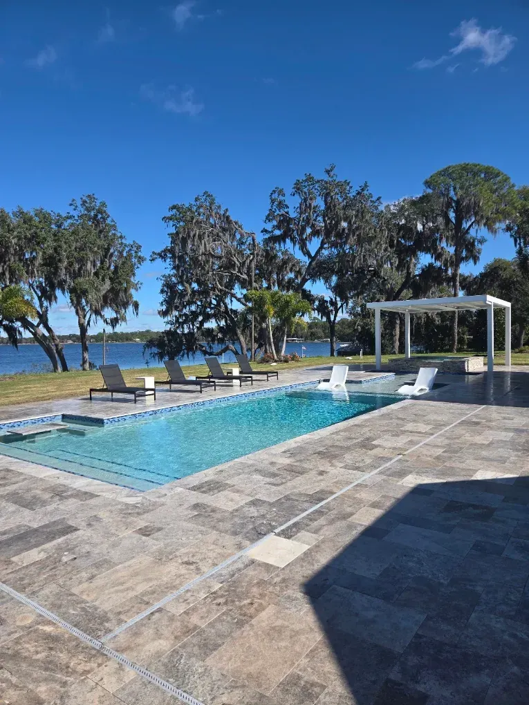 Outdoor pool beside the sea with lounge chairs, trees, and a white gazebo under a bright blue sky