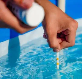 Hand holding a test strip over blue pool water, checking the water quality