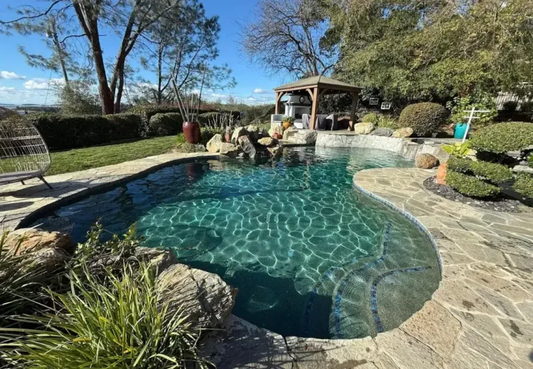 Curved backyard pool with turquoise water, stone deck, and a shaded seating area amid trees and landscaping