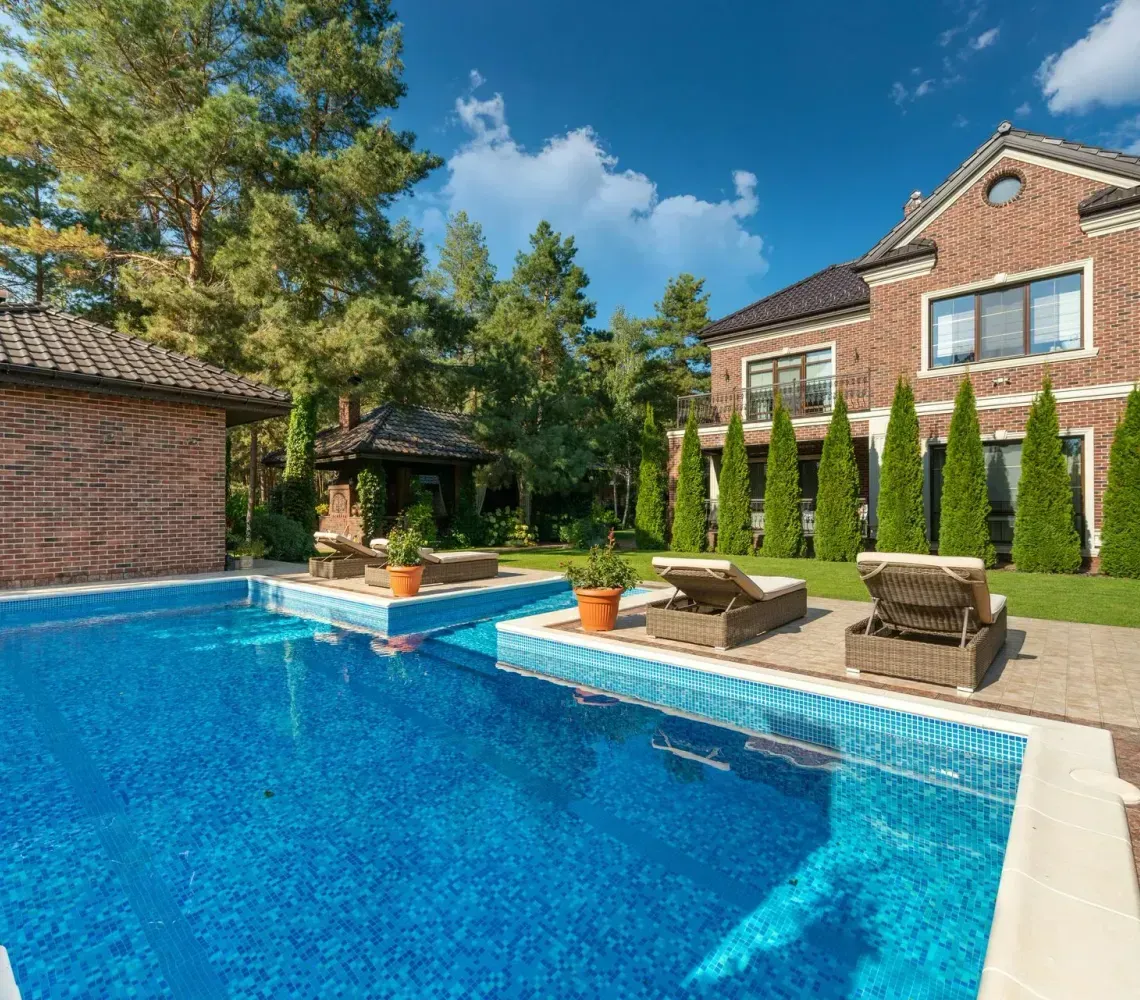 Backyard pool with blue water, stone patio, lounge chairs, and a brick house under sunny skies