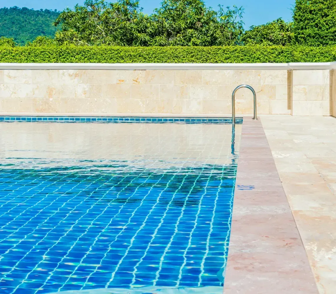 Blue-tiled swimming pool beside a stone deck with a clear blue sky and green trees beyond