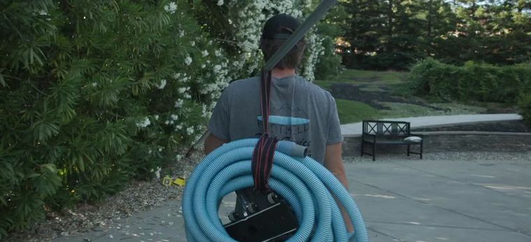 Person carrying a coiled blue hose on a wooded path near a driveway