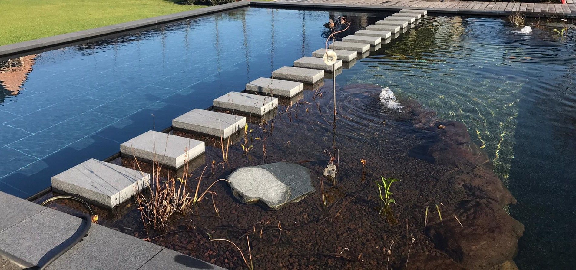 Stapstenen overbruggen een ondiepe vijver naast een smal blauw waterkanaal en een grasrijke oever.