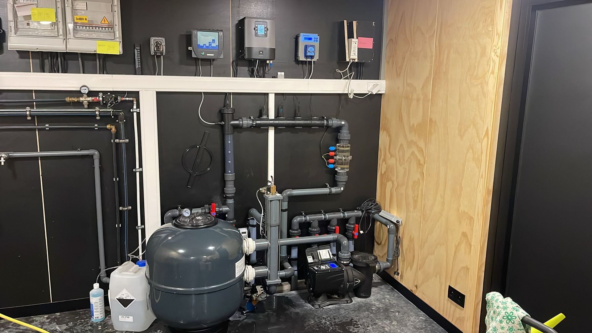Technische ruimte in de kelder met waterzuiveringstanks, leidingen, pompen en bedieningspanelen tegen een zwarte muur.
