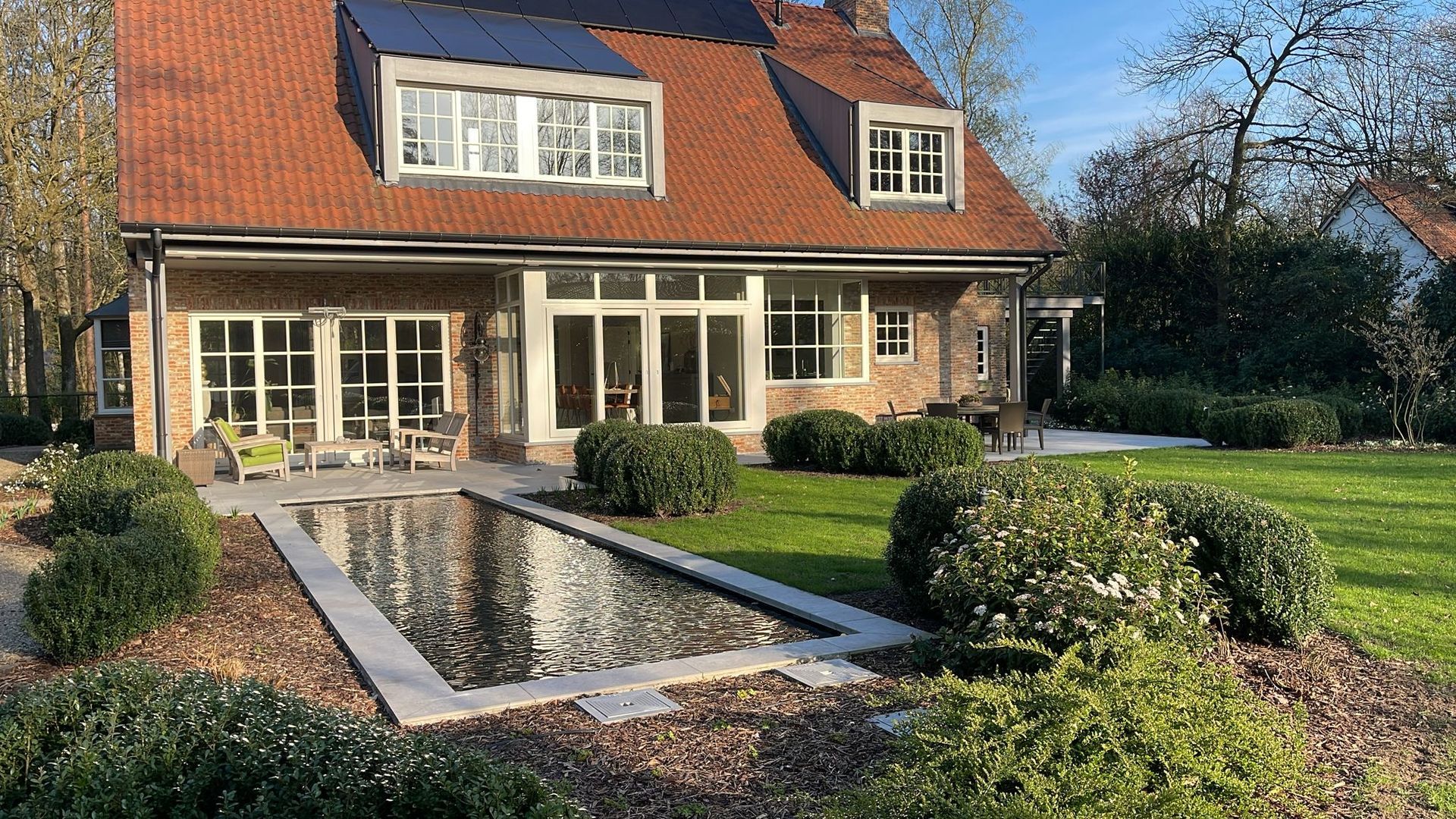 Bakstenen huis met rood dak, grote ramen en een aangelegde voortuin met een smalle vijver.