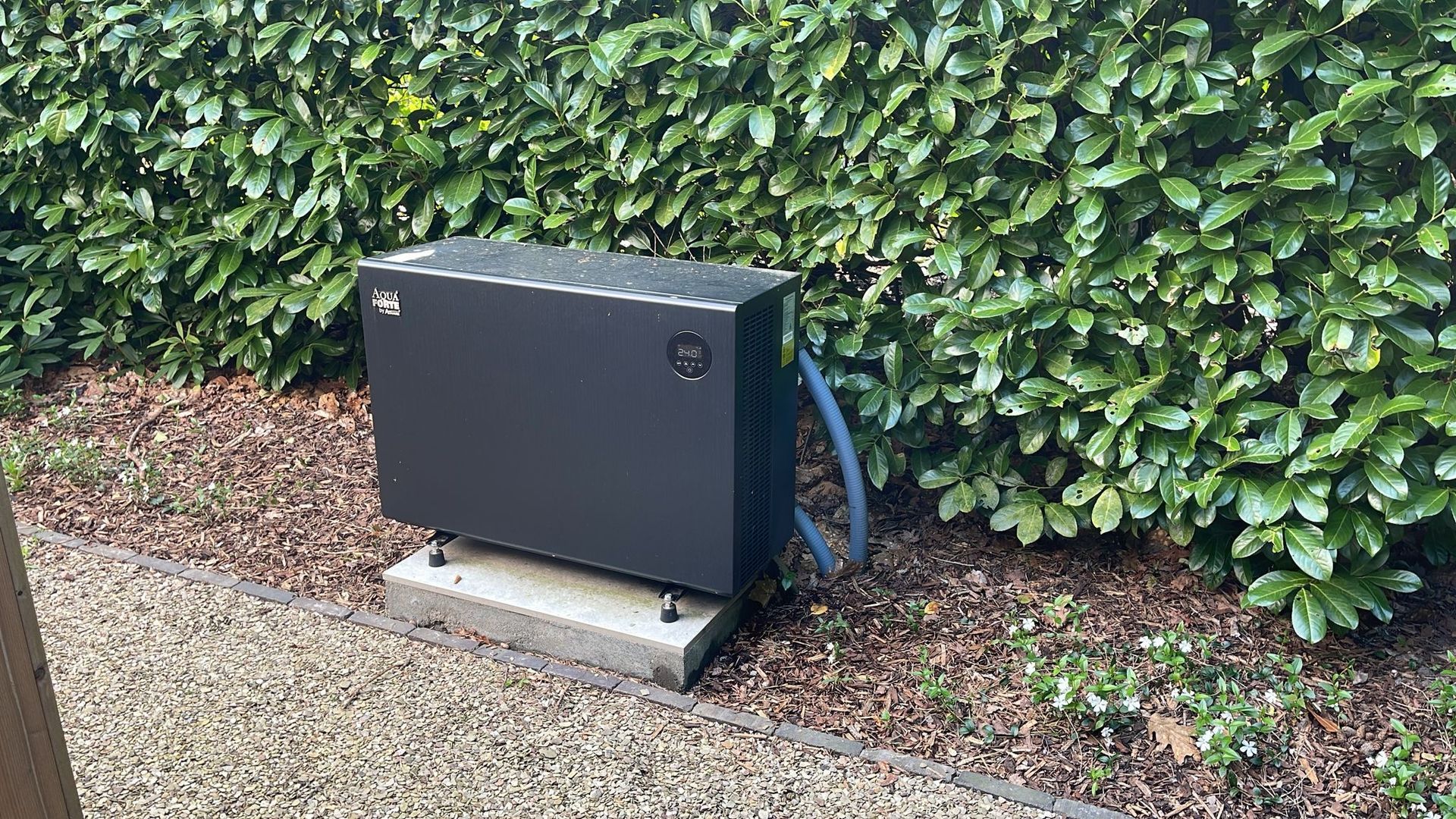 Zwarte elektriciteitskast naast een heg op een grindpad, met een blauwe slang eraan bevestigd.