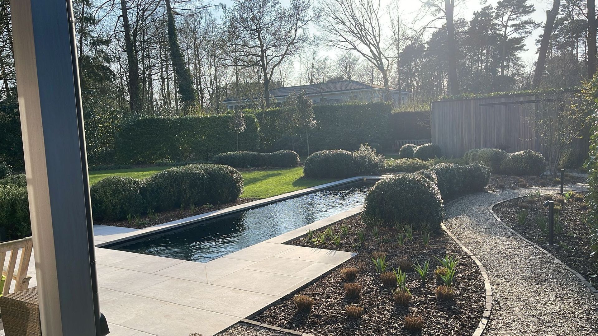 Een zonnige achtertuin met een rechthoekig reflectiebad, een grindpad en strak gesnoeide hagen.
