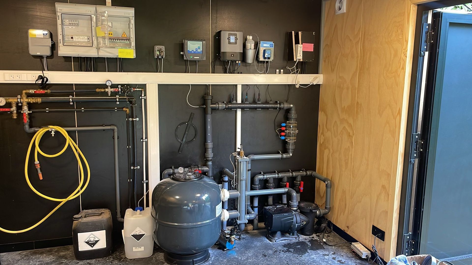 Technische ruimte met tanks, leidingen, bedieningspanelen en opgerolde slangen tegen onafgewerkte muren.