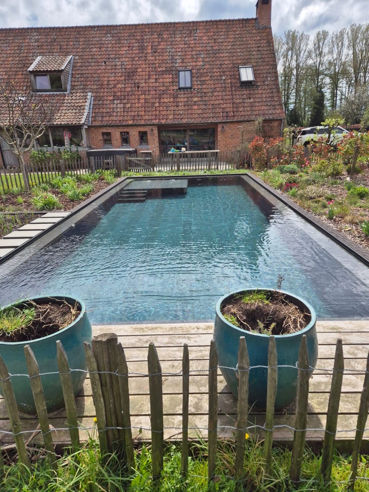 Rechthoekige zwarte vijver met stenen rand in een aangelegde tuin, begrensd door een houten schutting en hagen.