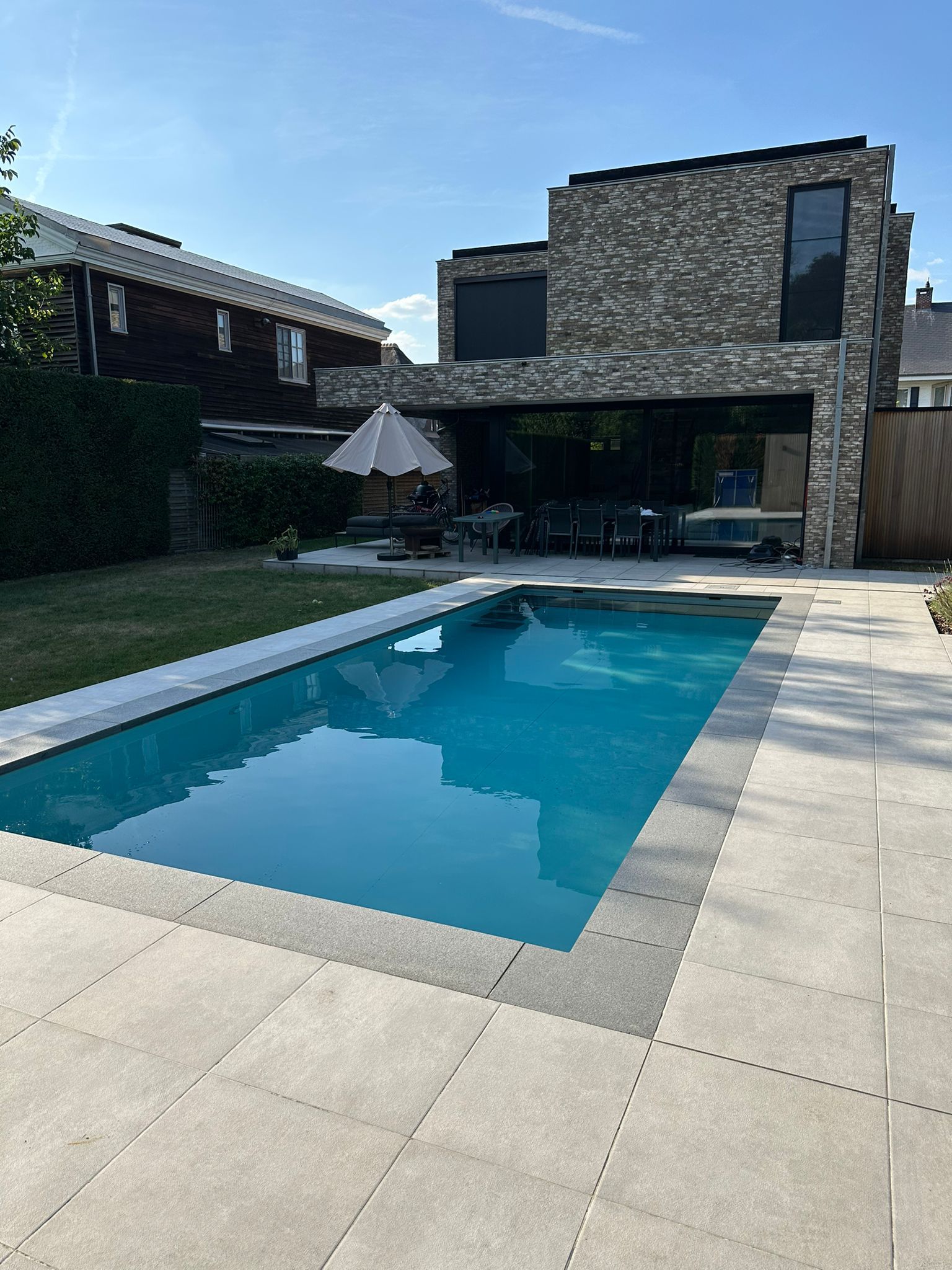 Moderne achtertuin met een rechthoekig zwembad naast een stenen huis en een parasol.