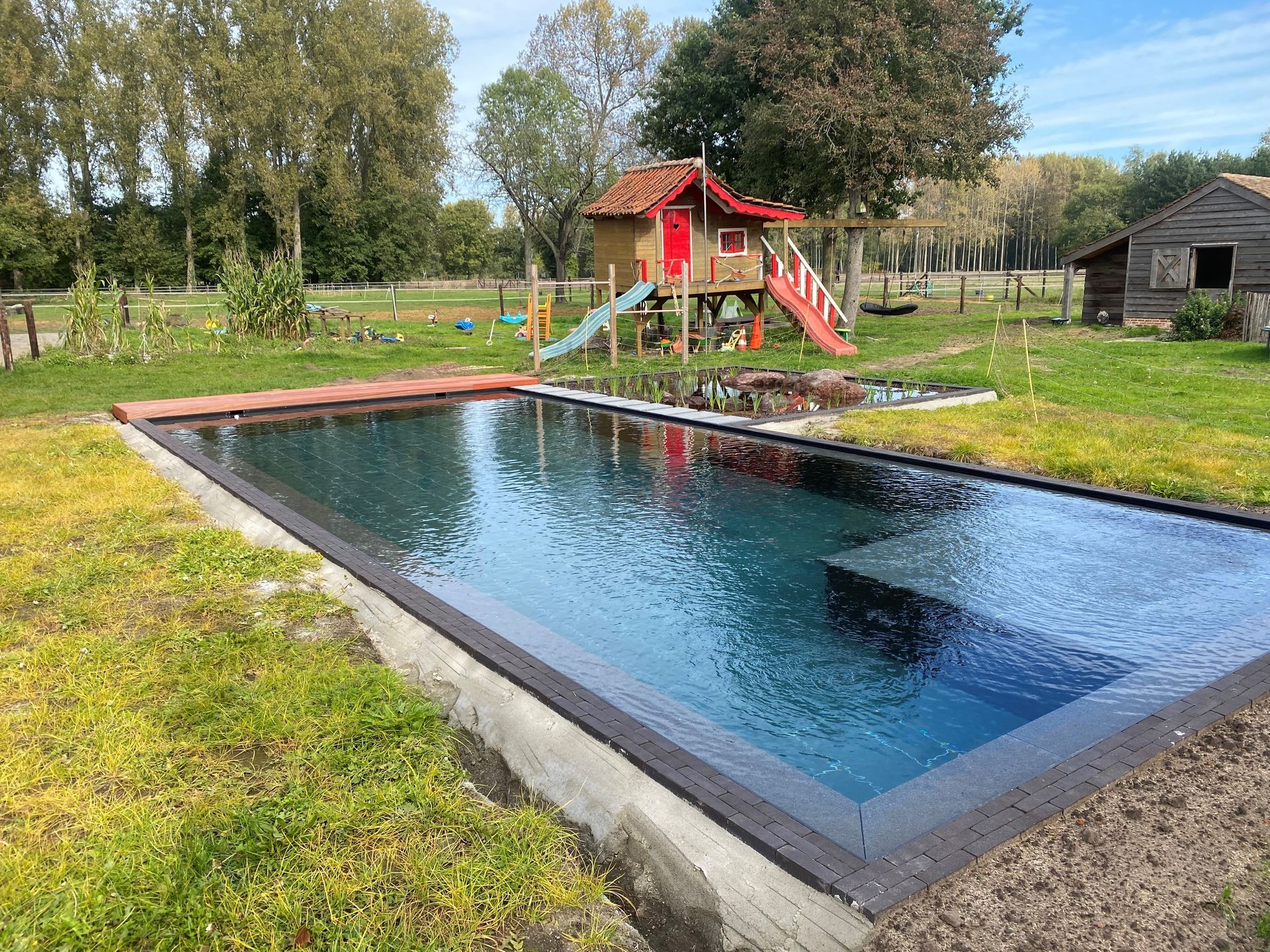 Een groot zwembad omgeven door gras en bomen met een speeltuin op de achtergrond.