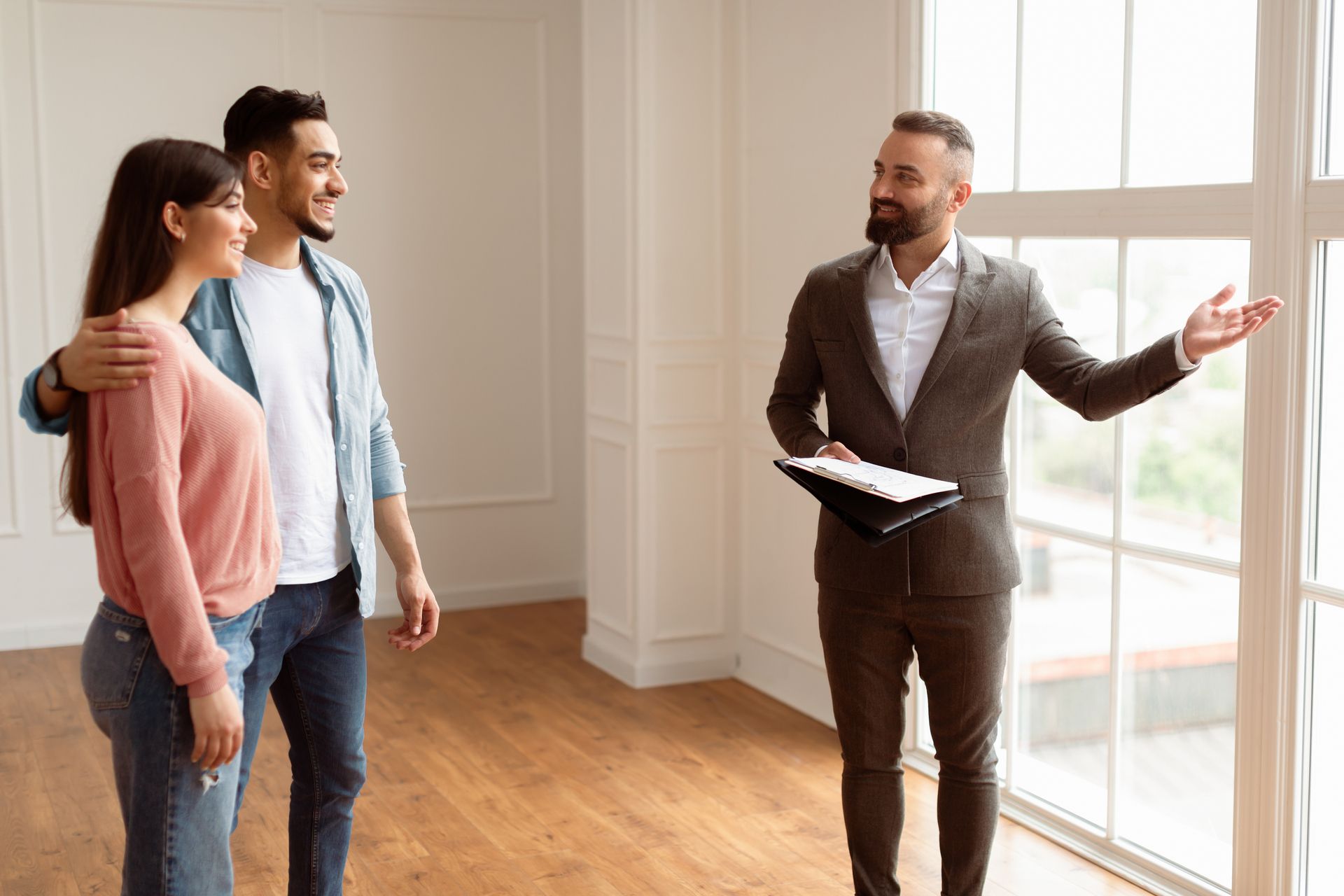 Real estate agent showing a young couple a property, pointing at a large window.