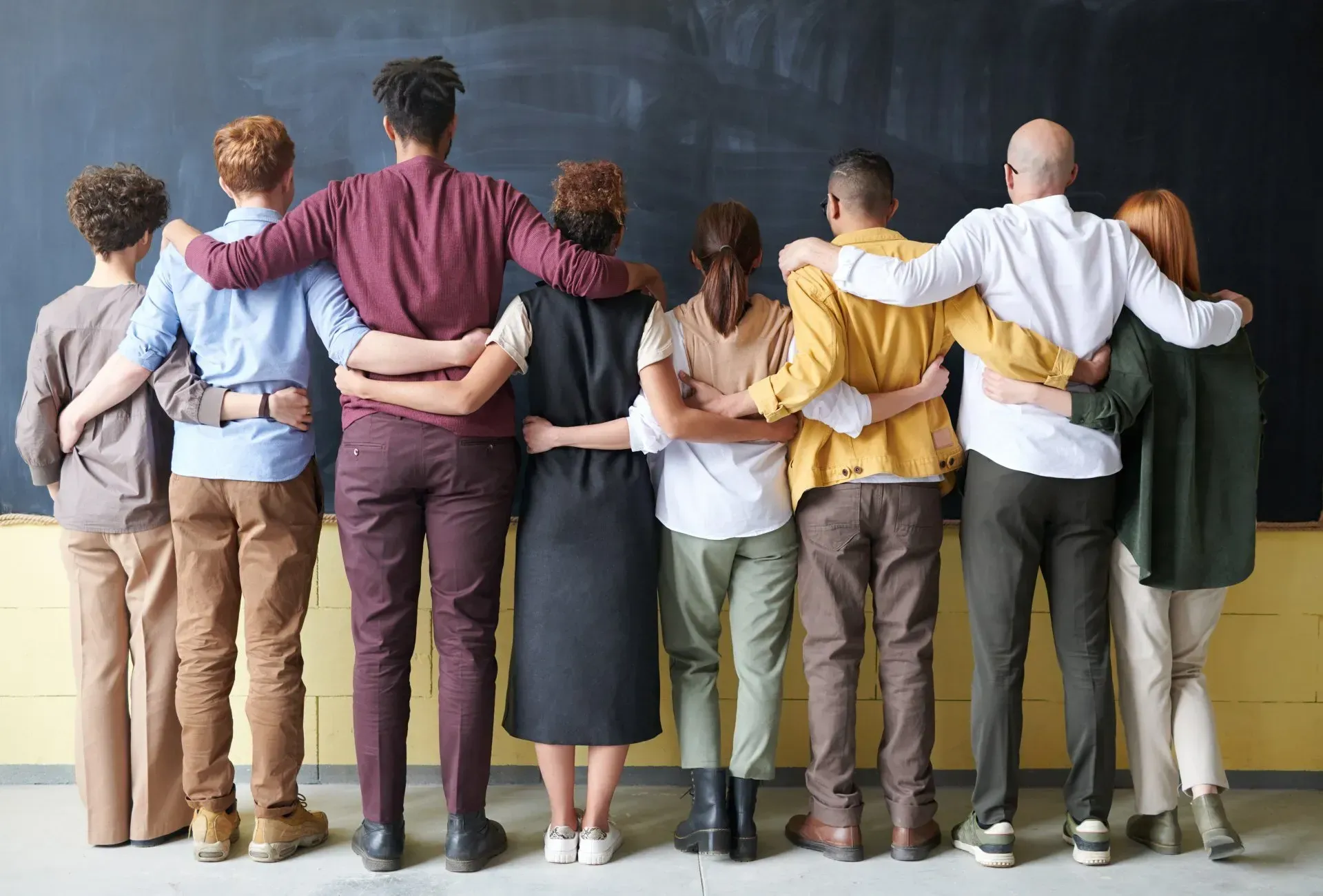Group of diverse people with arms around each other, standing in front of a blackboard.