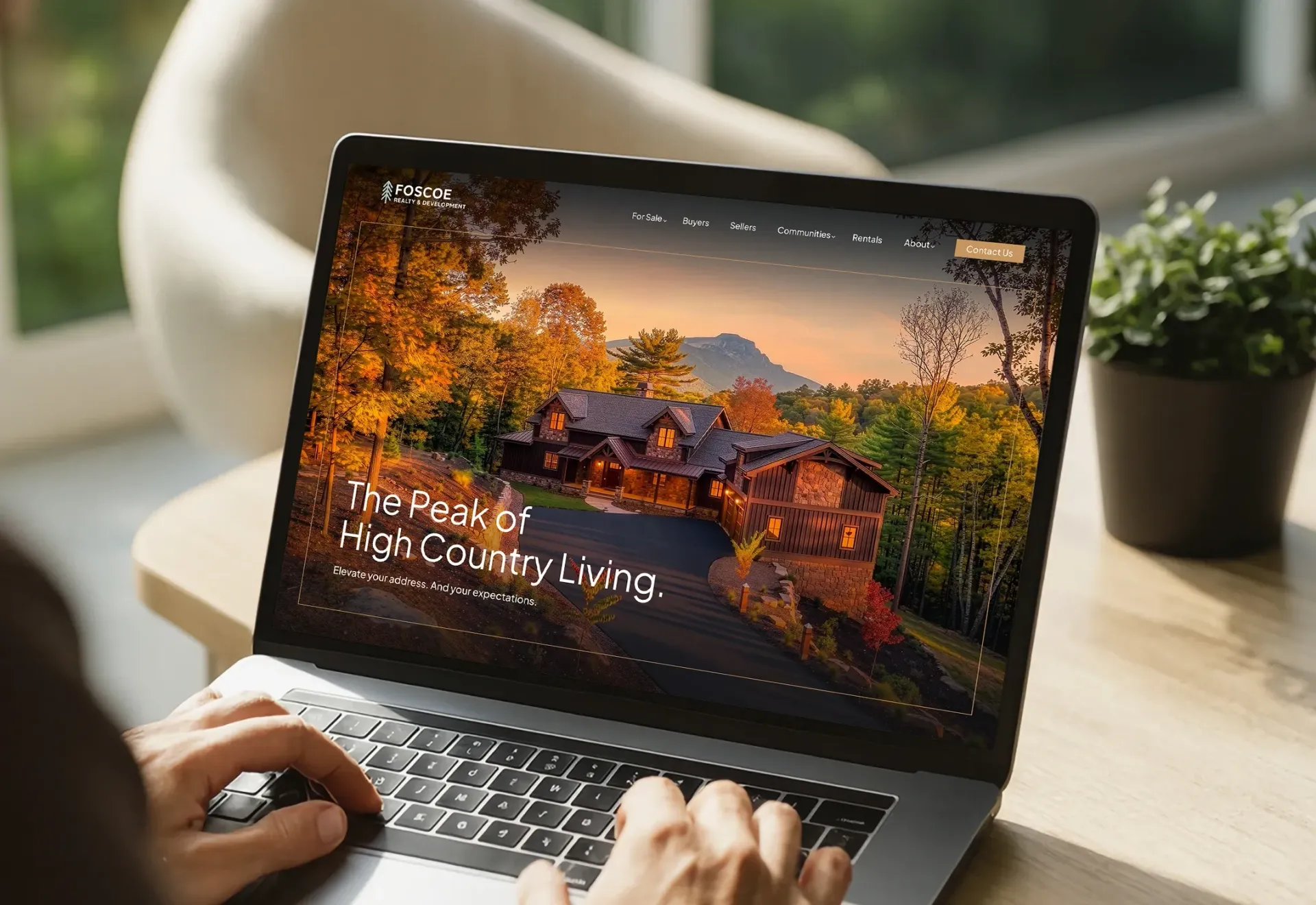 Hands typing on a laptop showing a scenic autumn website hero section on a desk by a potted plant
