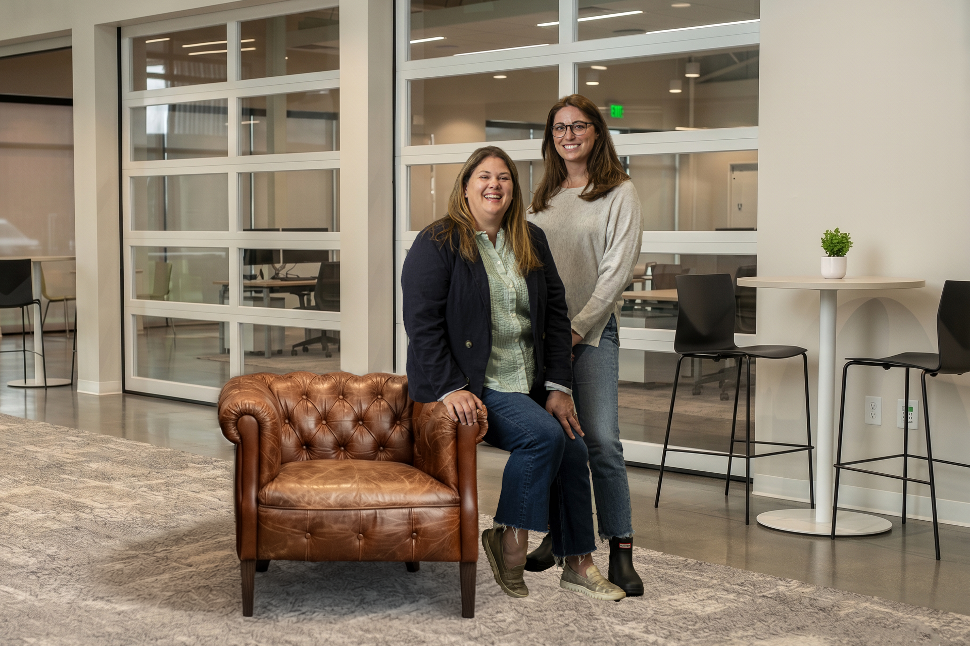 Two people standing in a modern office lounge with glass walls and a brown leather couch.