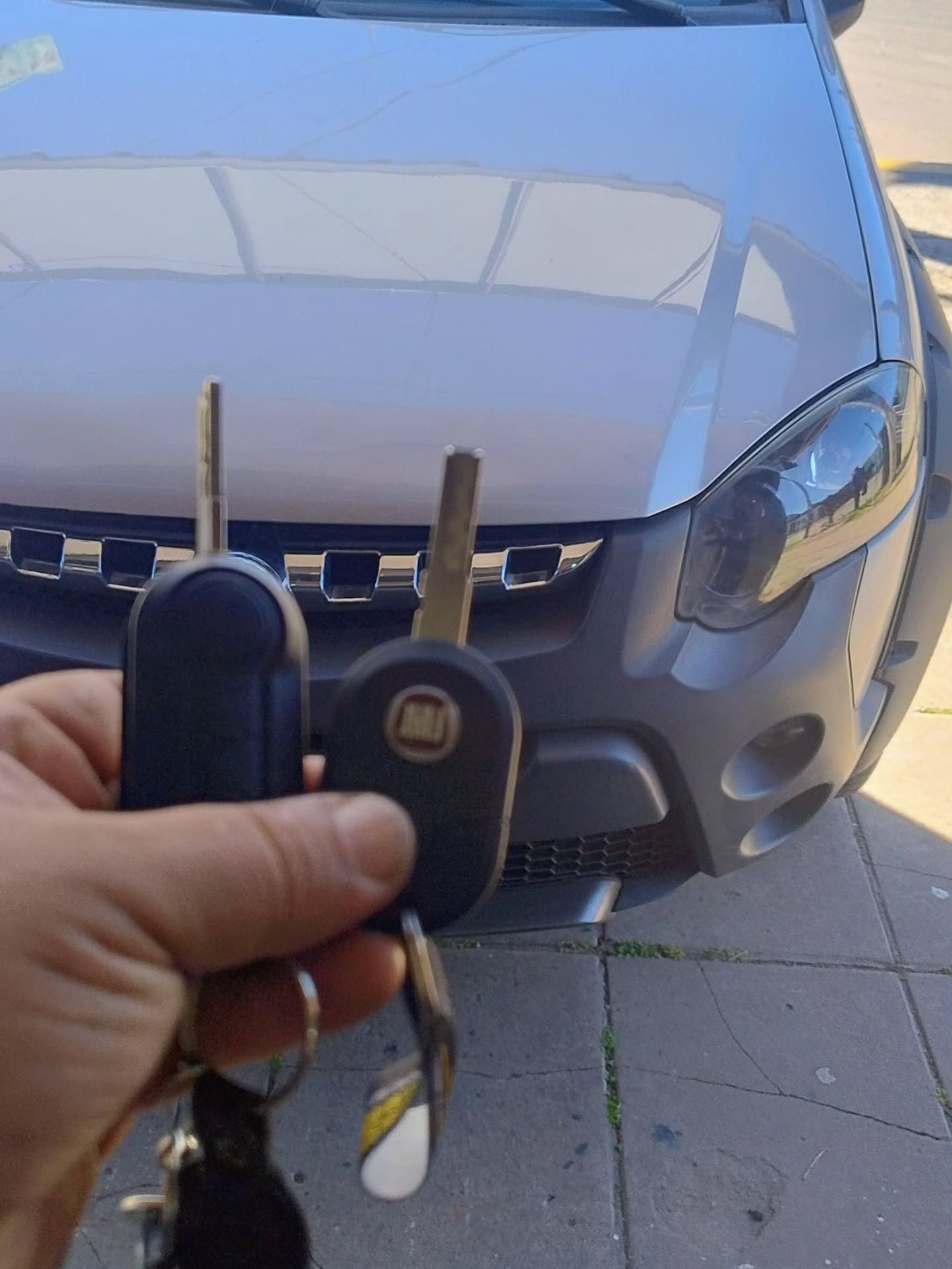 Mano sosteniendo dos llaves de coche delante de un Fiat plateado.