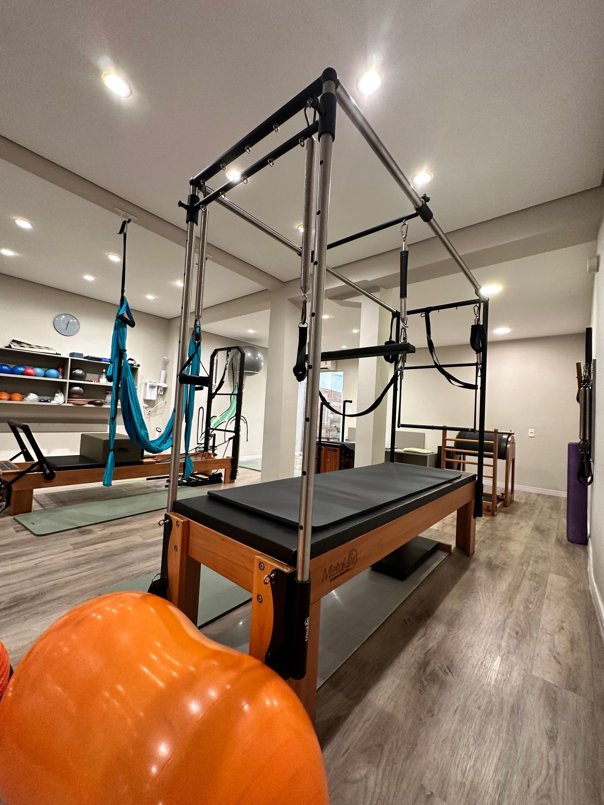 Estúdio de Pilates com equipamento Reformer, piso de madeira e uma grande bola de exercícios laranja em primeiro plano.