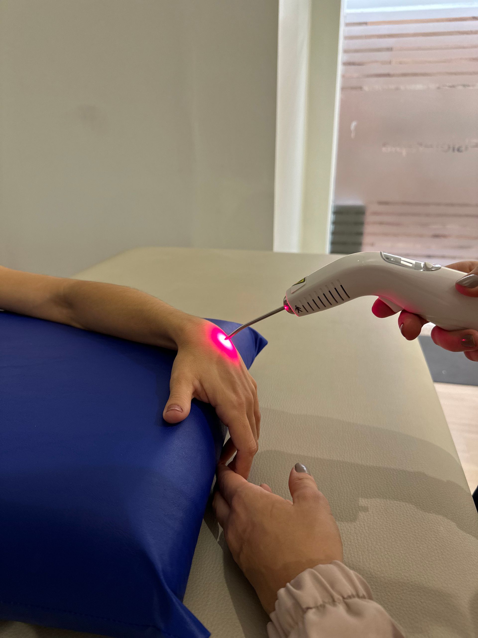 Mão examinando o antebraço com um termômetro de luz vermelha ao lado de uma almofada azul sobre uma mesa.