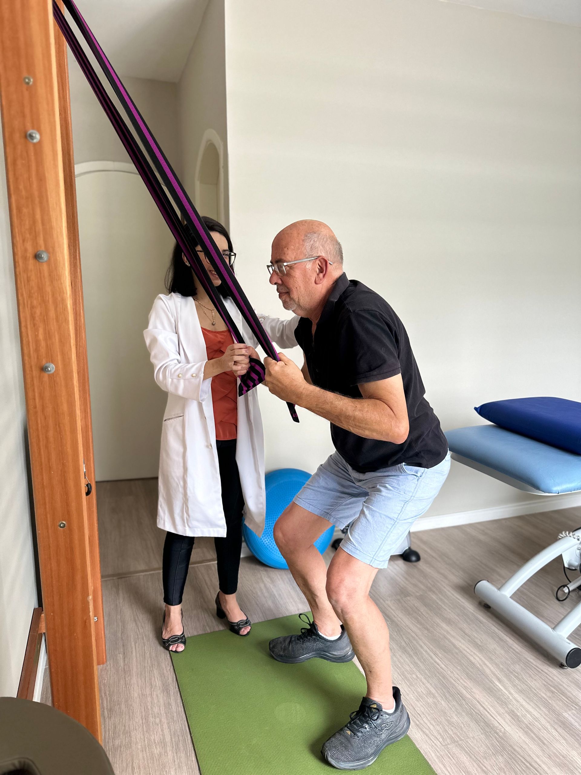 Exercício de fisioterapia com faixas de resistência em uma sala de clínica; paciente e fisioterapeuta trabalhando juntos.