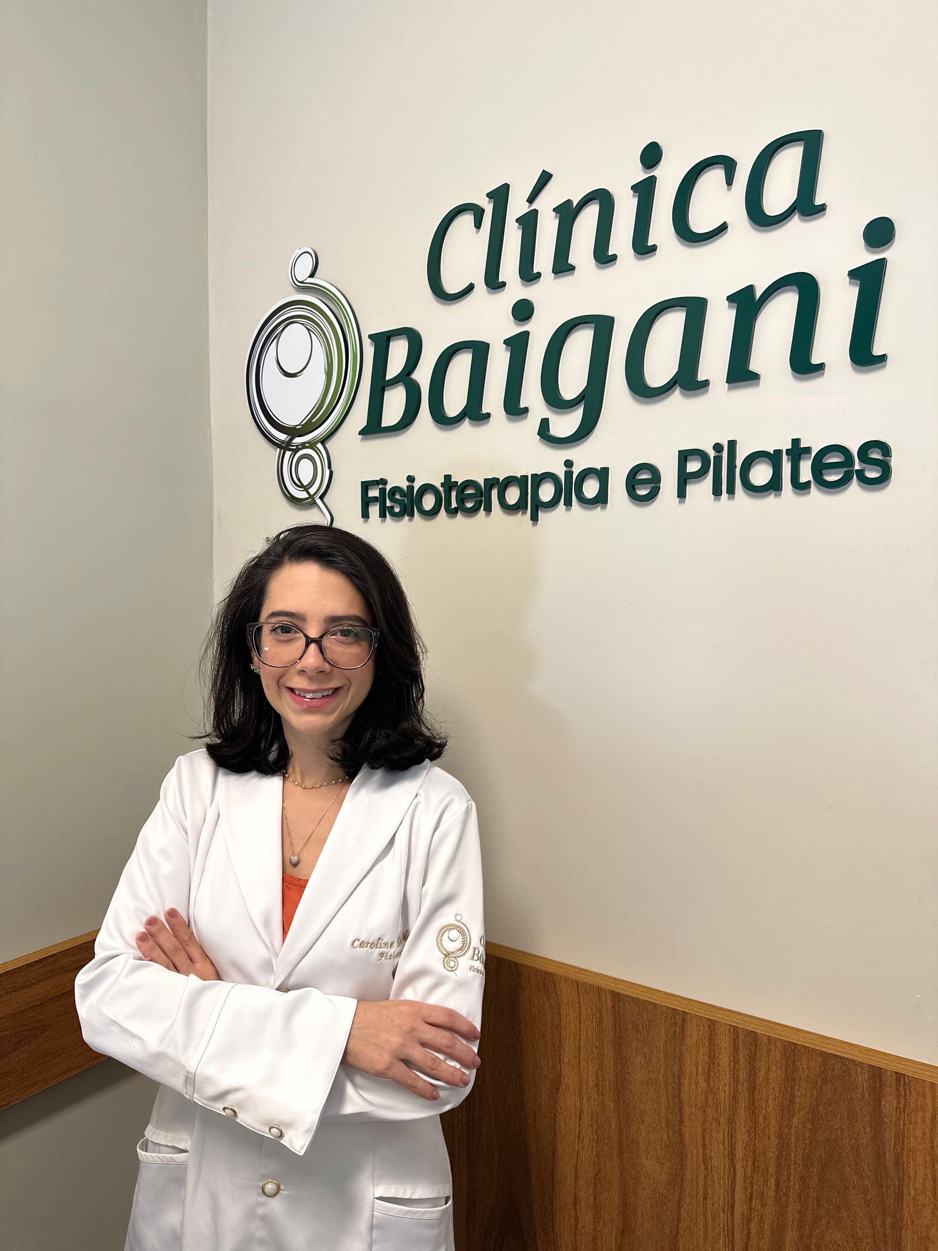 Mulher de óculos em pé numa clínica, com os braços cruzados, ao lado de uma placa com os dizeres Clínica Baigani
