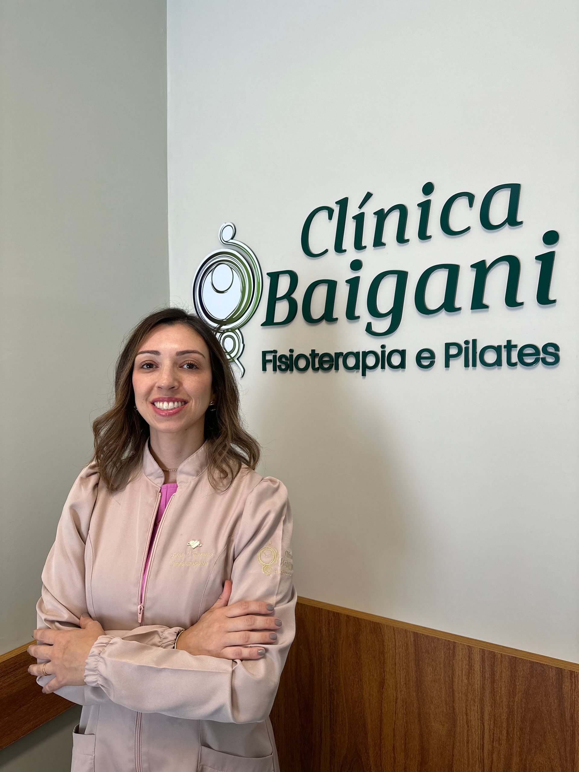 Mulher em pé ao lado da placa da Clínica Baigani no corredor da clínica, vestindo um blazer bege.