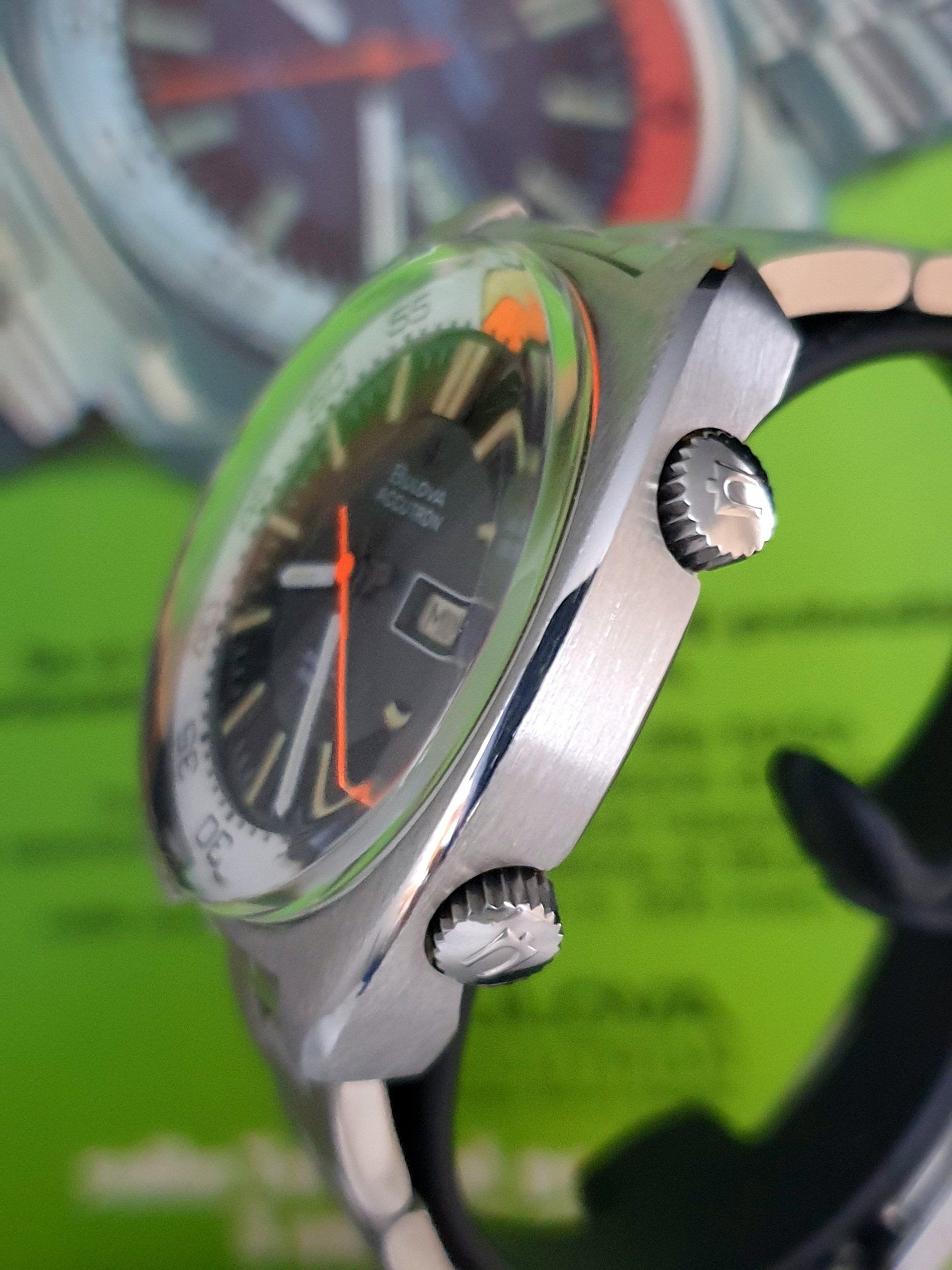 Close-up of a silver watch with a black face and orange hands on a green background