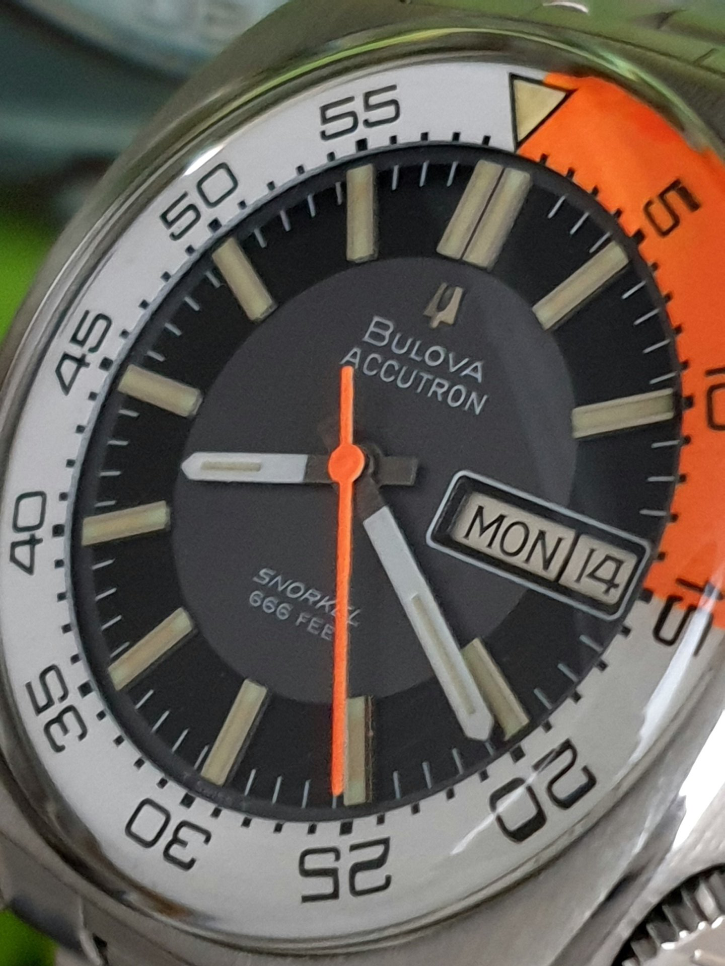 Close-up of a Bulova Accutron wristwatch with black dial, silver bezel, orange accents, and day date display