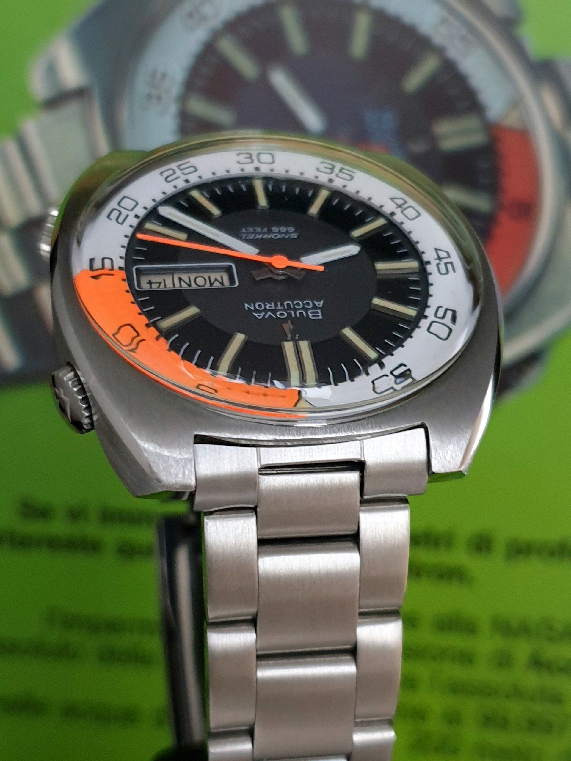 Silver wristwatch with orange accent and black face on a green background