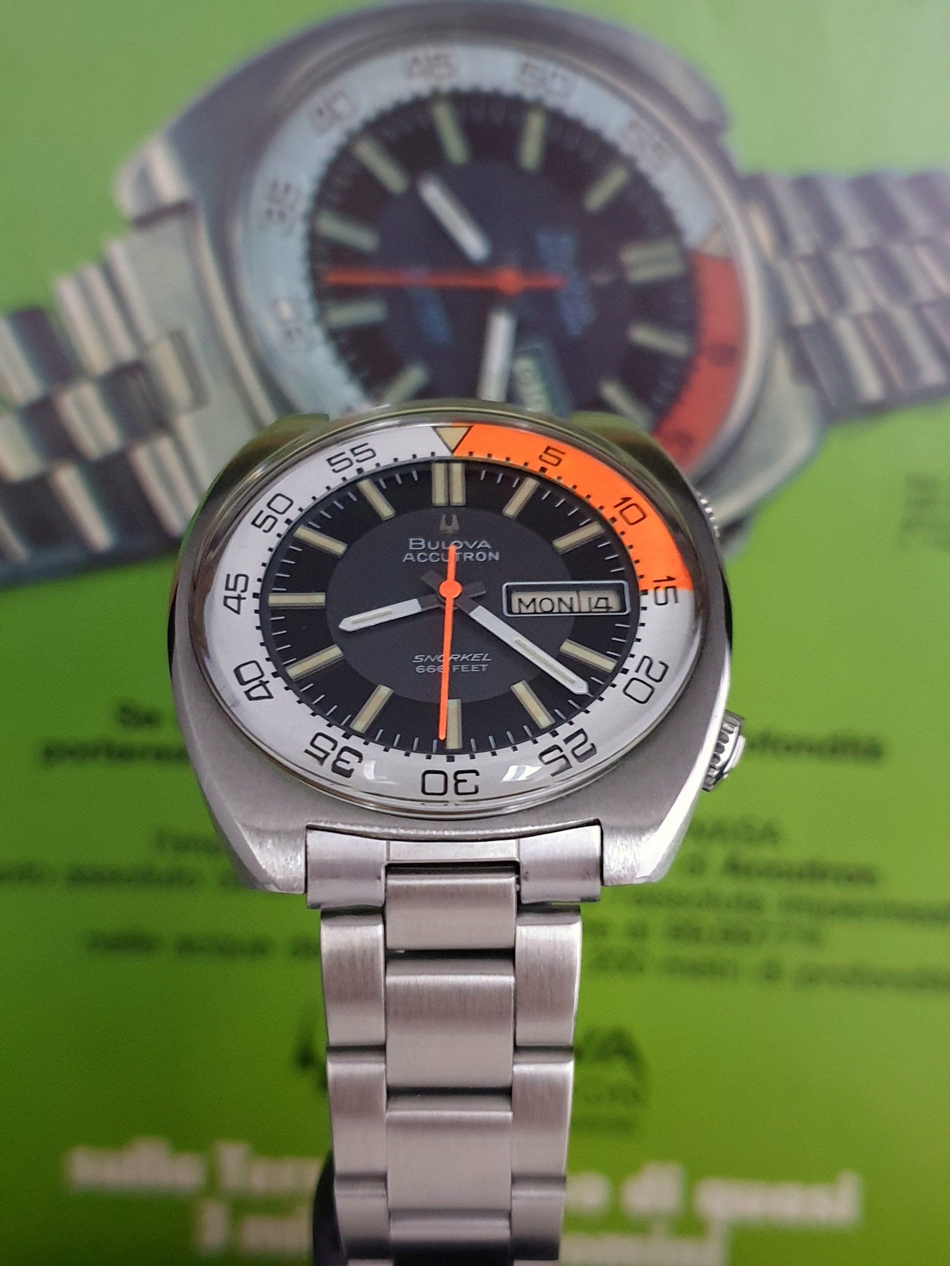 Stainless steel analog wristwatch with black and orange dial, shown on a green background.