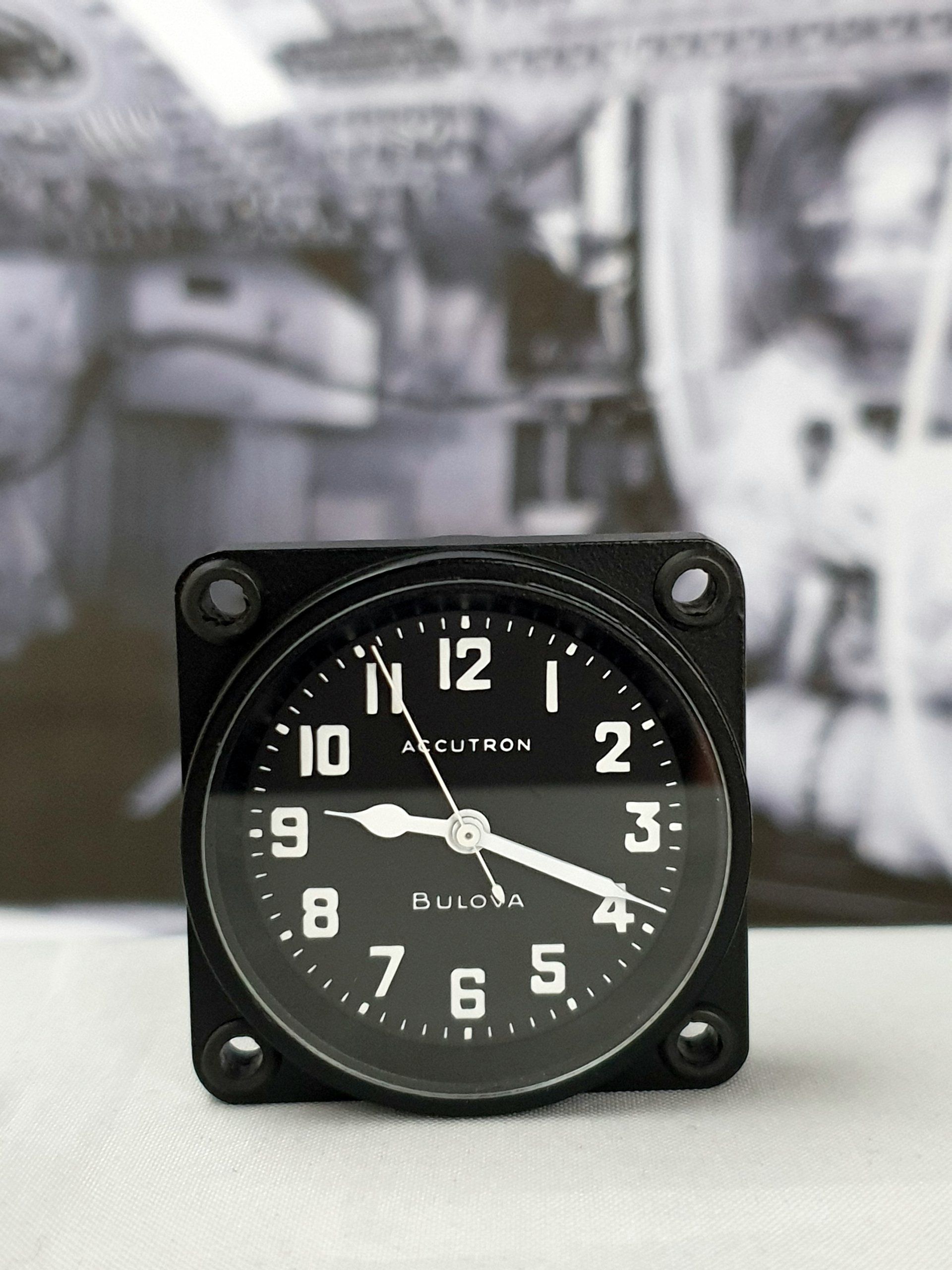 Black square analog clock with white numbers on a blurred industrial background