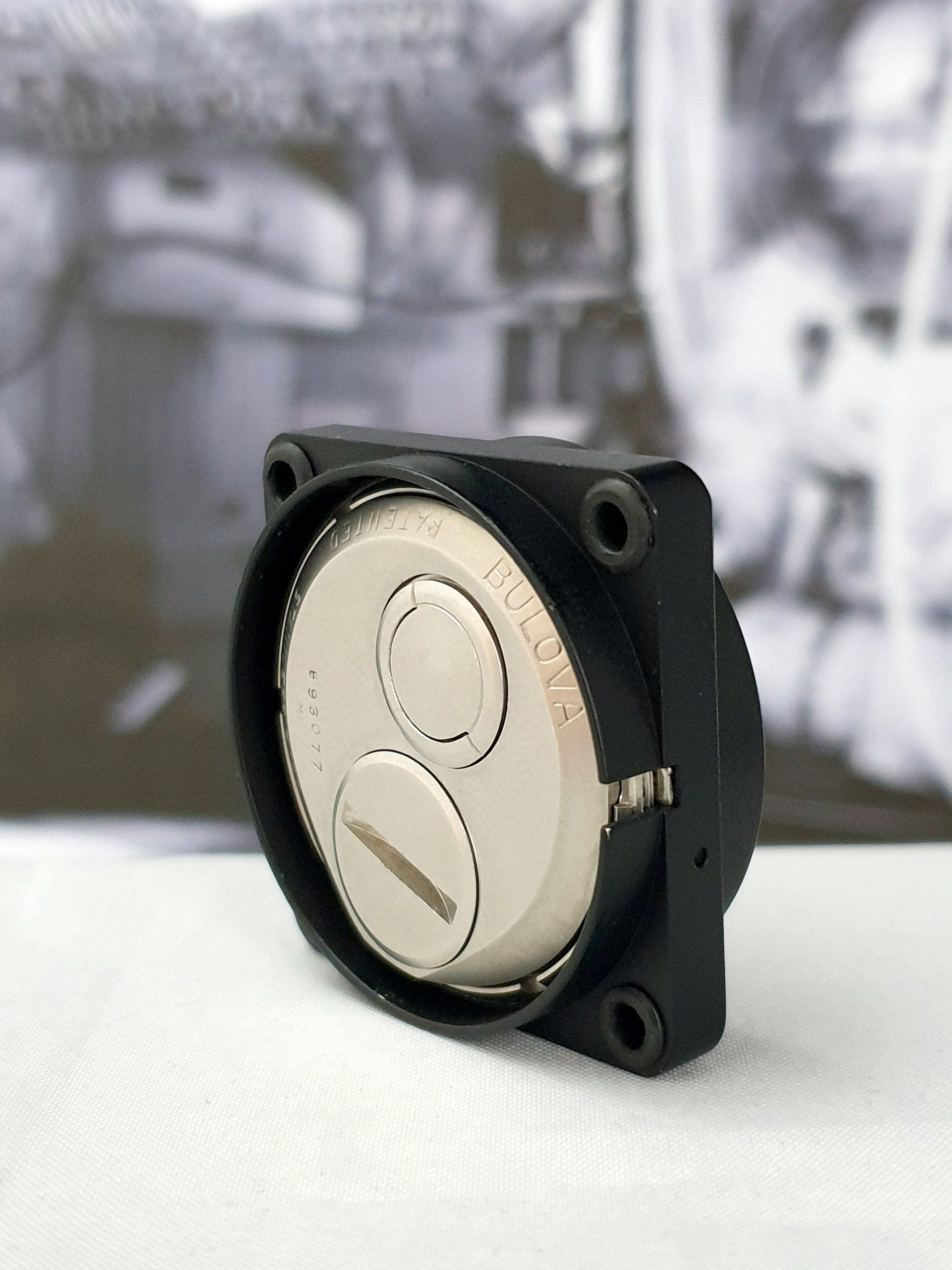Black square device with a silver circular face, shown on a tabletop against a blurred background