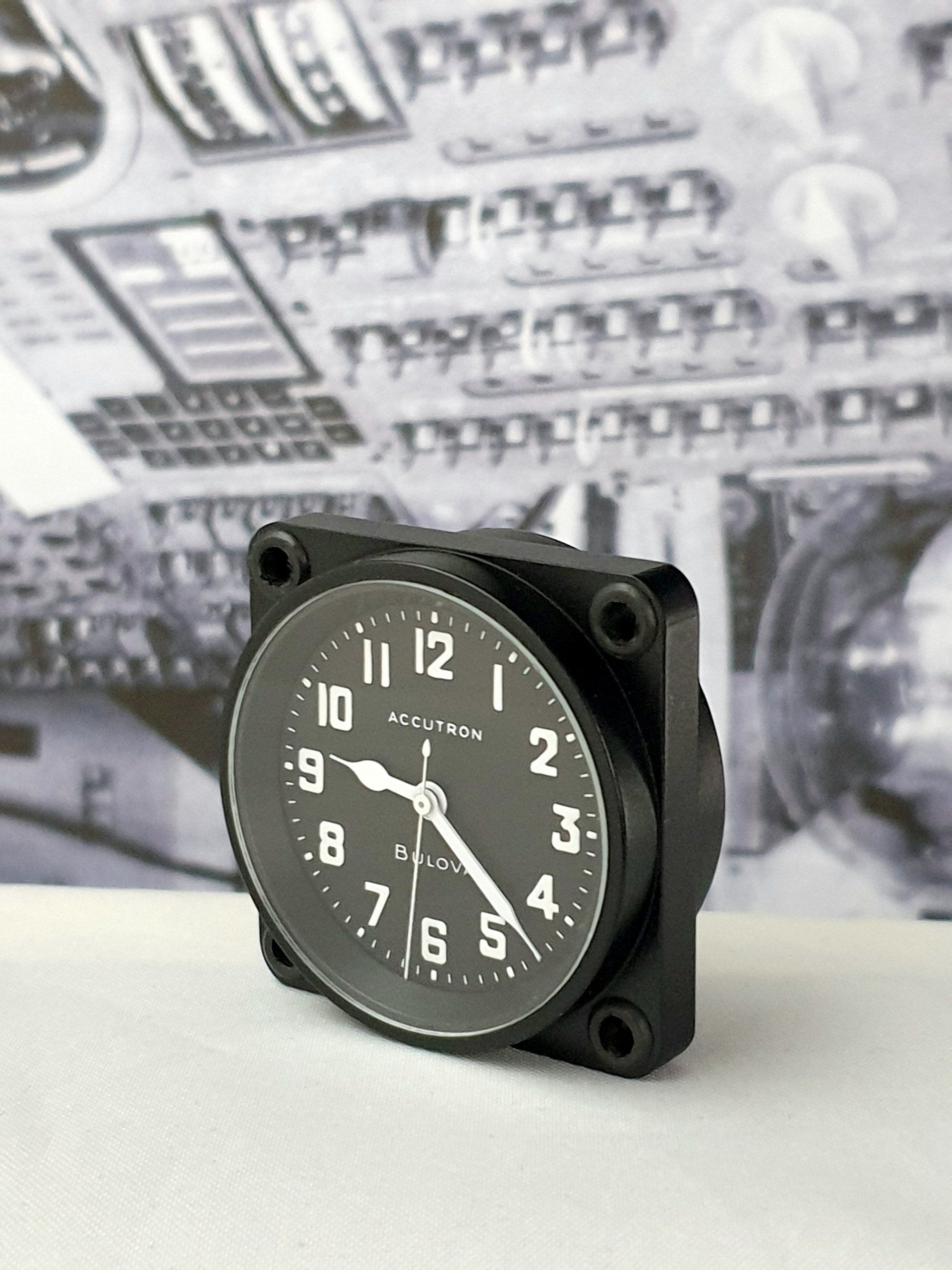 Black analog clock with white numbers on a black panel, set on a white surface before blurred equipment.