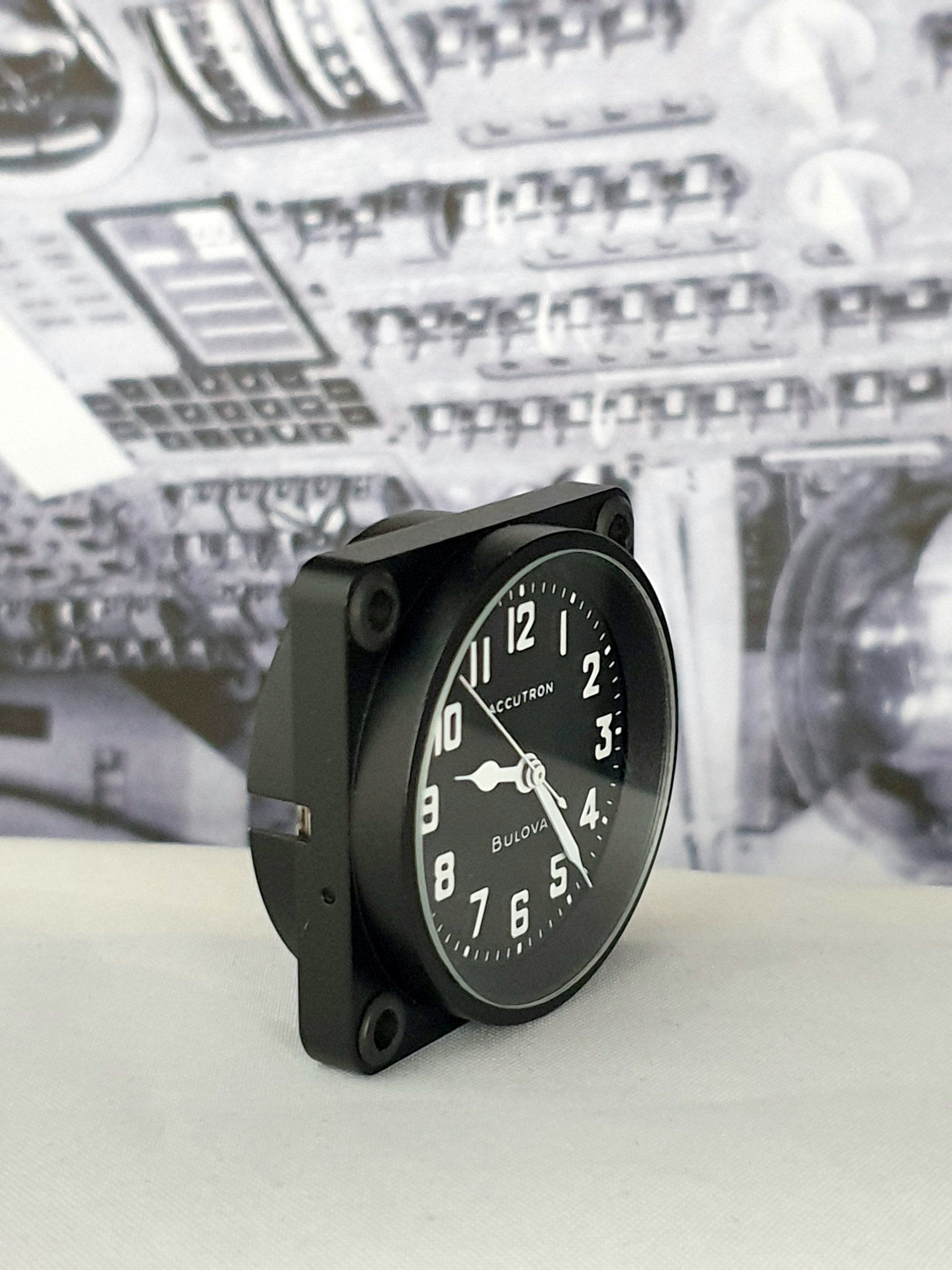 Black analog clock resting on a white surface, with blurred circuit board background.