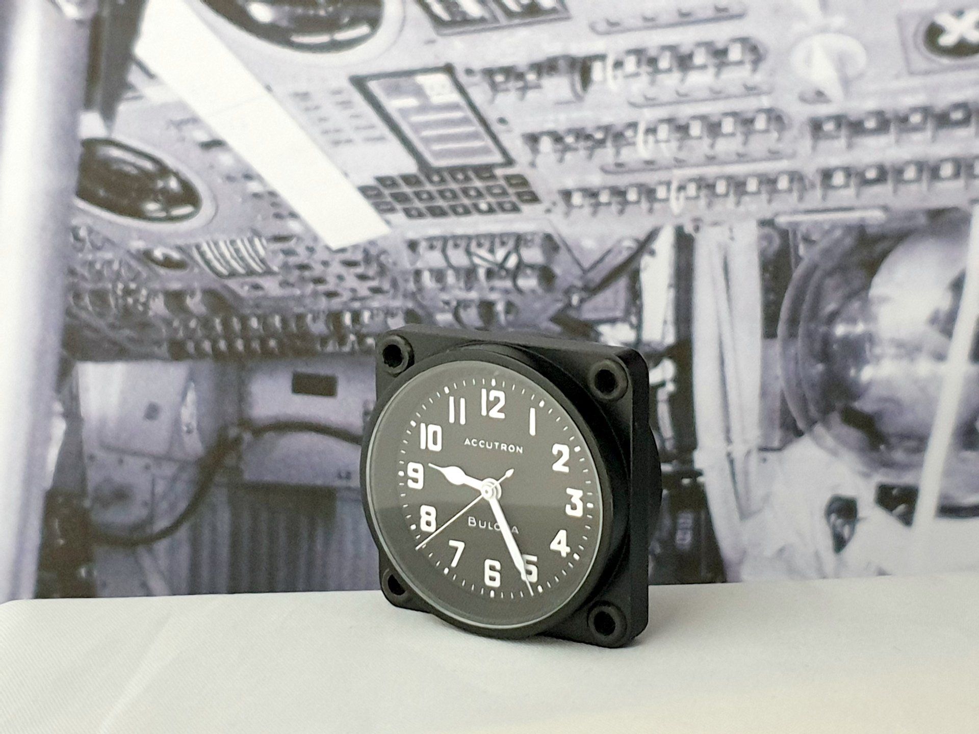 Small black analog clock on a white surface with a gray mechanical background