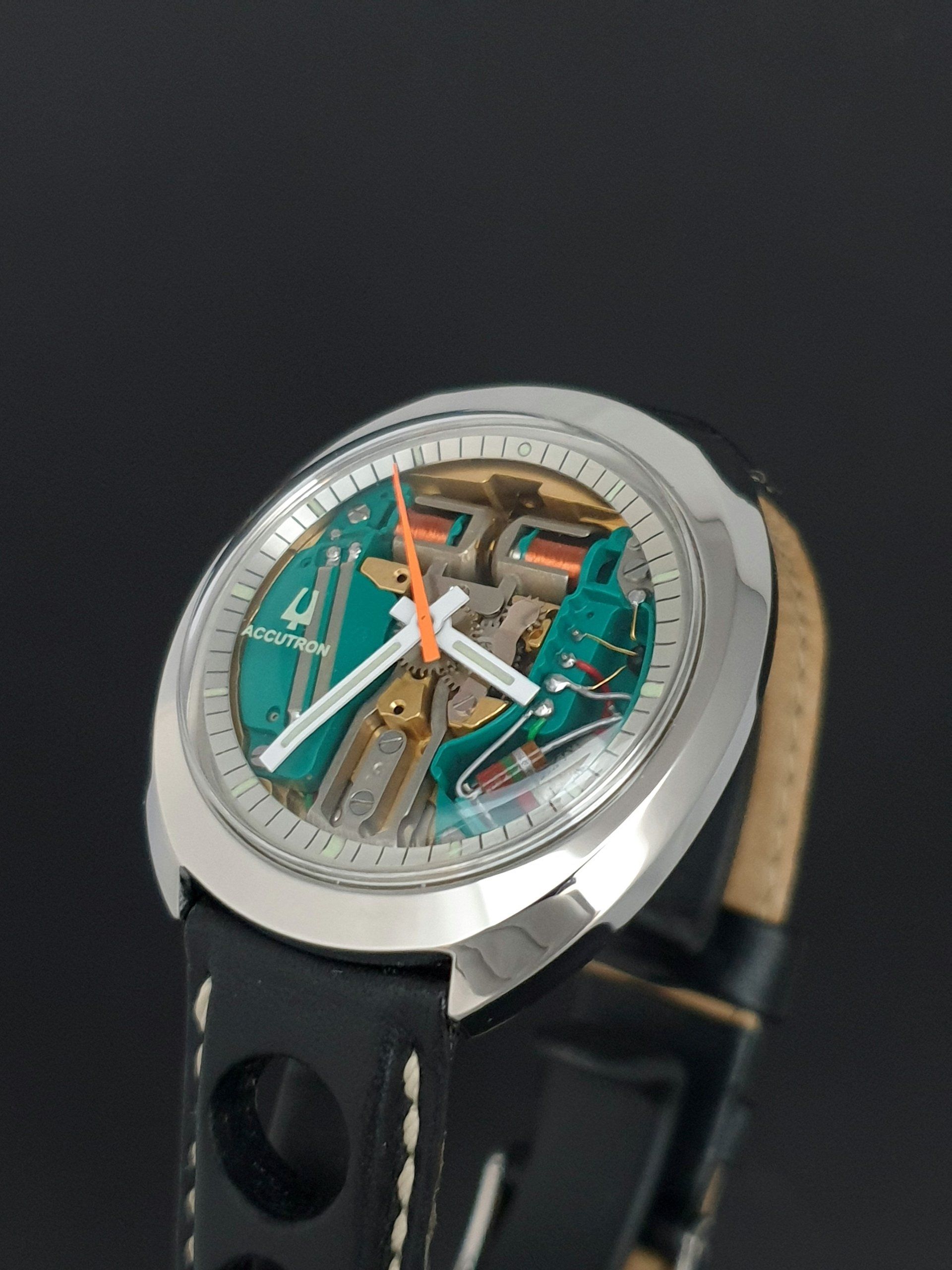 Close-up of a silver watch with a teal skeleton dial and black strap on a dark background