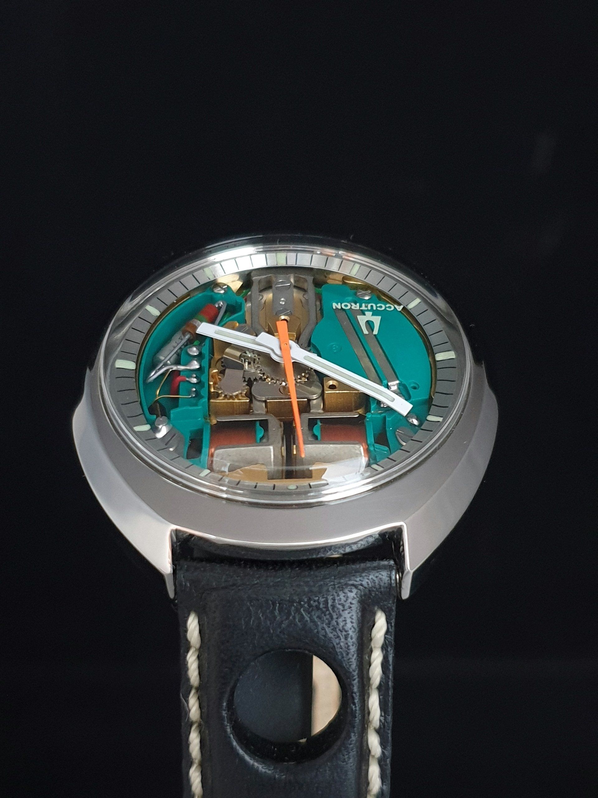 Silver wristwatch with a black leather strap and teal skeleton dial on a dark background