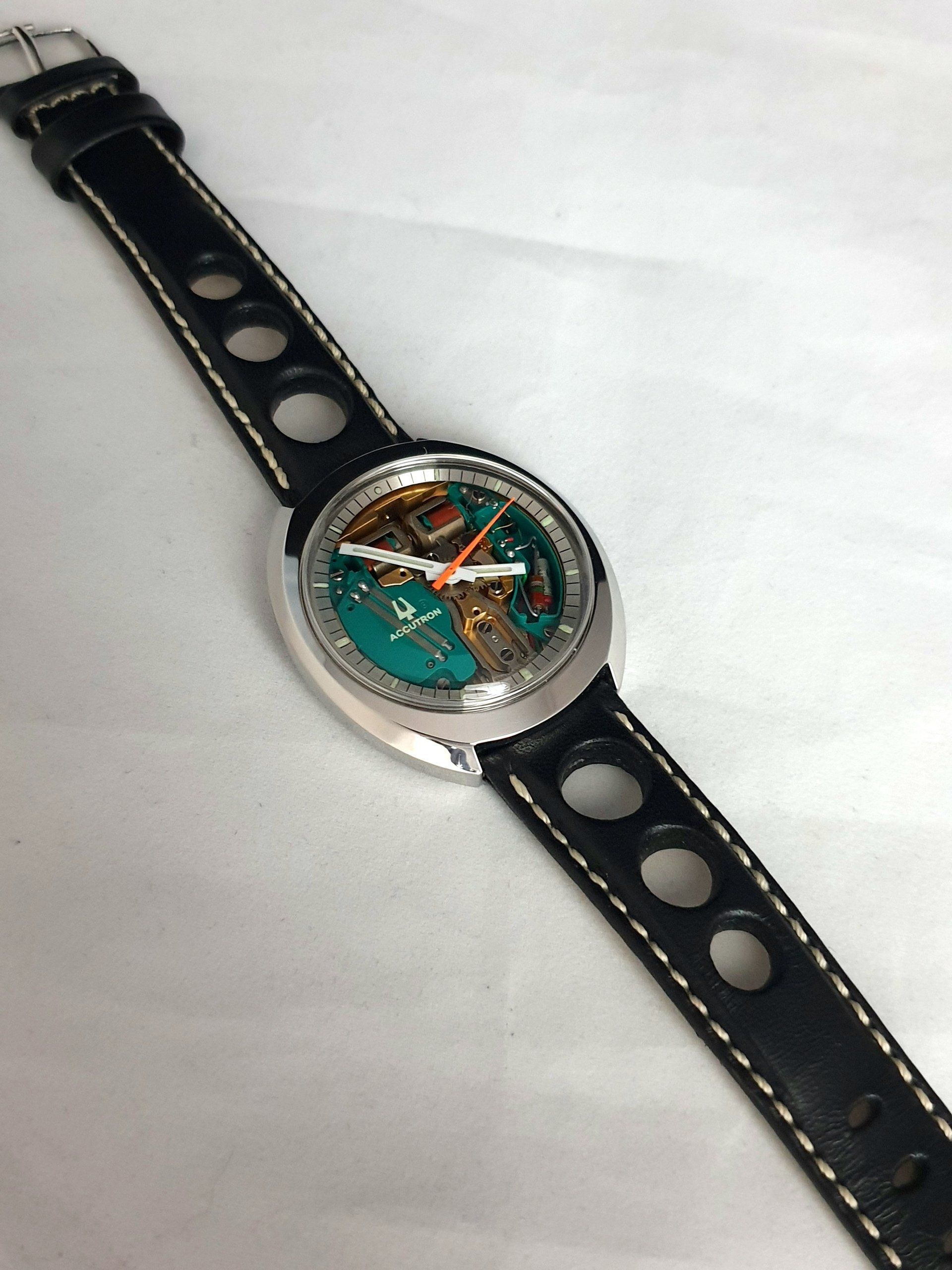Round wristwatch with black perforated strap and teal dial on a white background