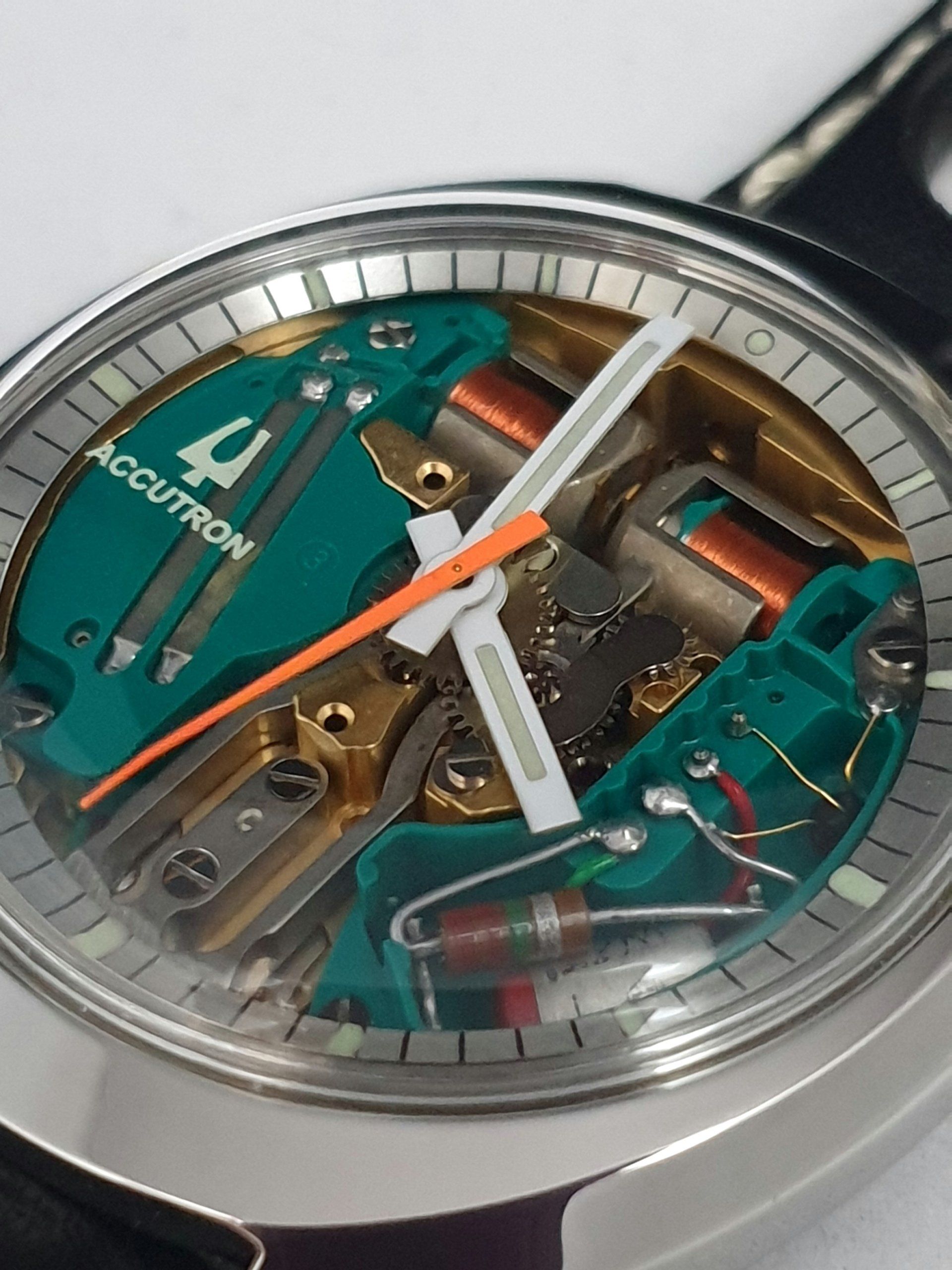 Close-up of a transparent wristwatch with teal components, orange seconds hand, and visible internal mechanics