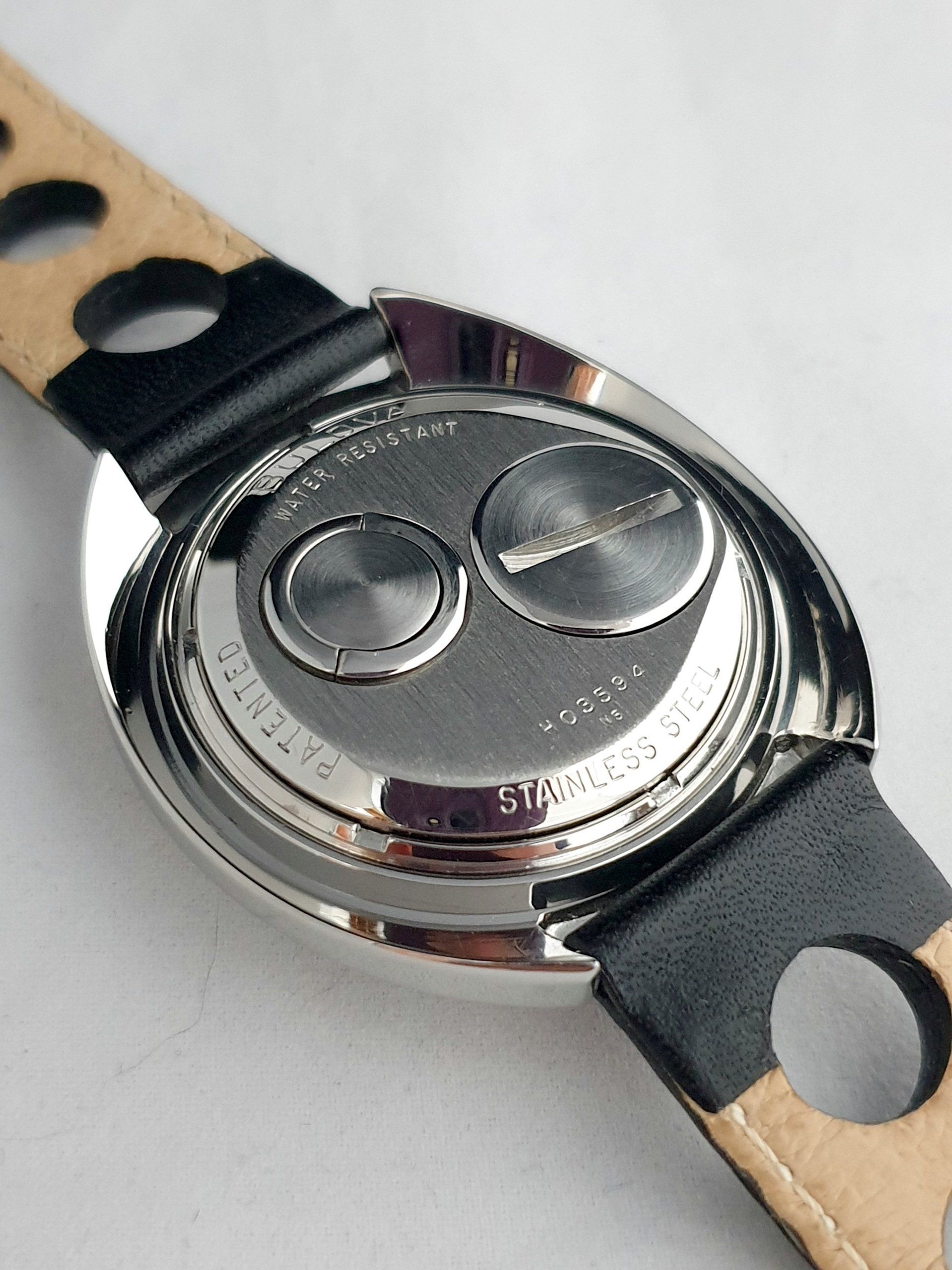 Back of a silver wristwatch with a black strap and tan leather underside, showing two crowns/buttons.