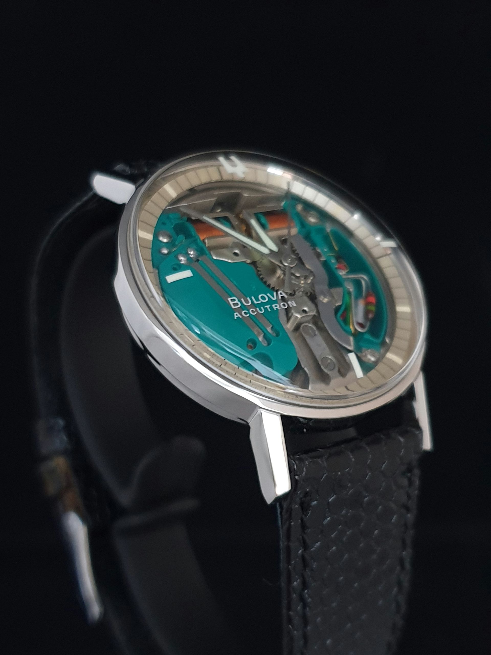 Close-up of a silver skeleton wristwatch with a teal dial and black textured leather strap