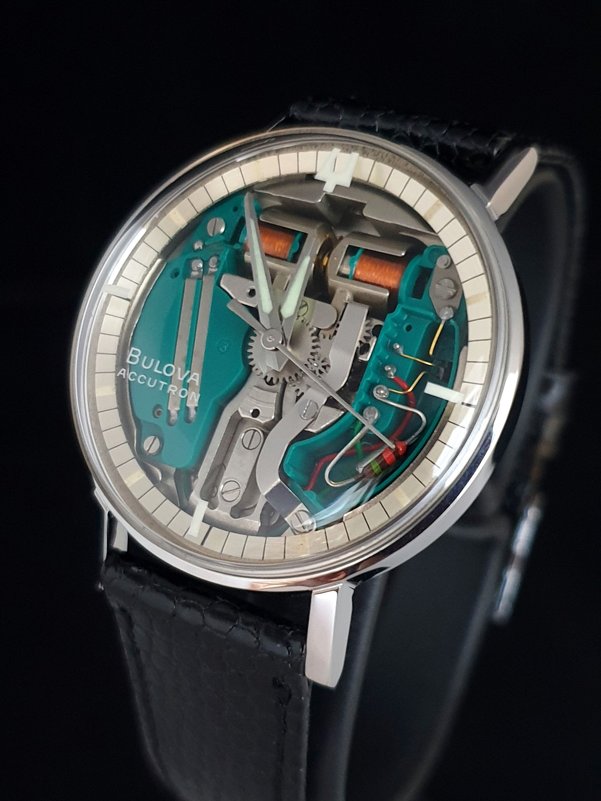 Skeleton wristwatch with silver case, teal dial accents, and black leather strap on a dark background