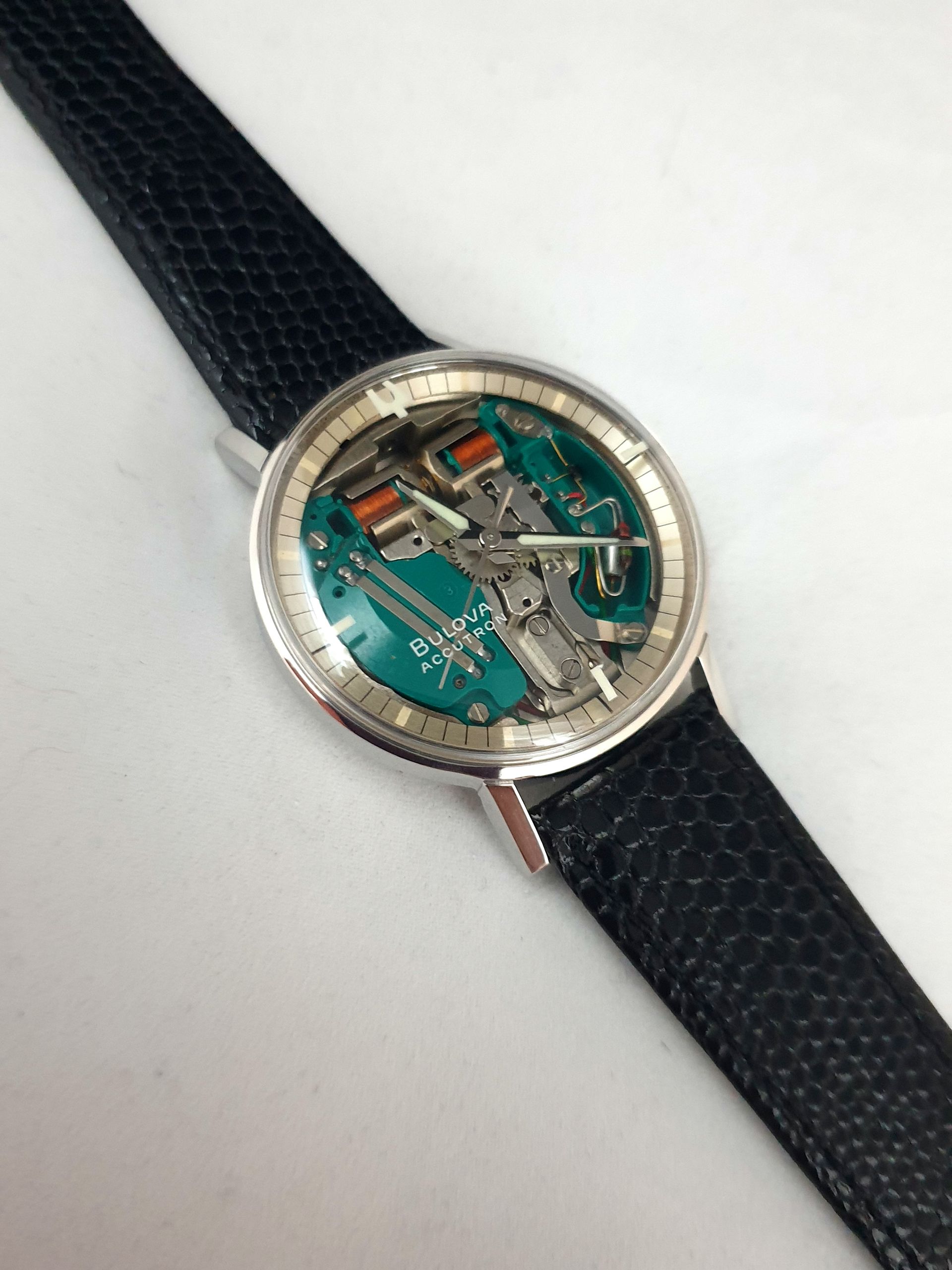 Close-up of a silver wristwatch with a teal skeleton dial and black textured strap on a white background
