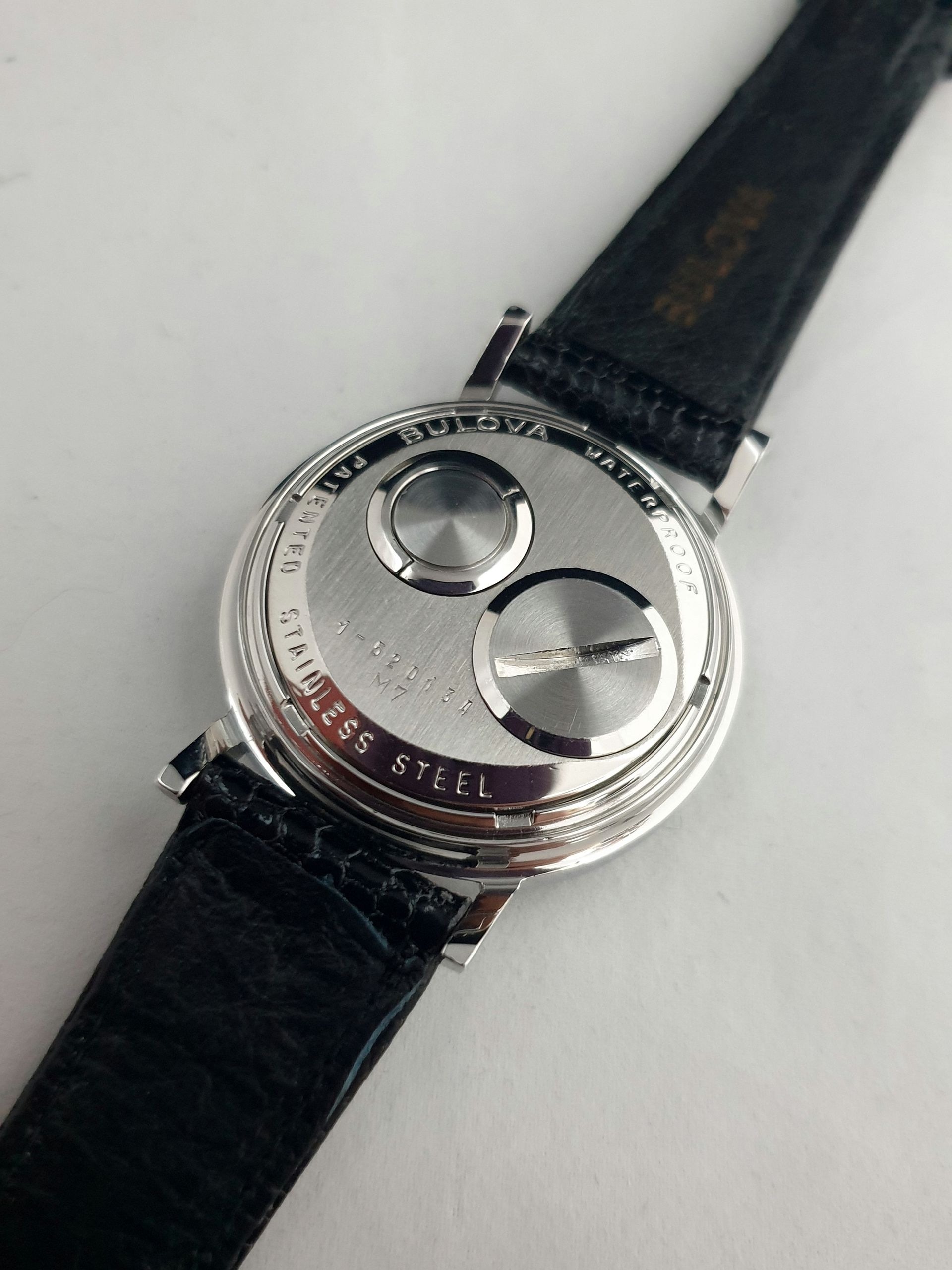 Silver watch case back with two round covers on a black leather strap.