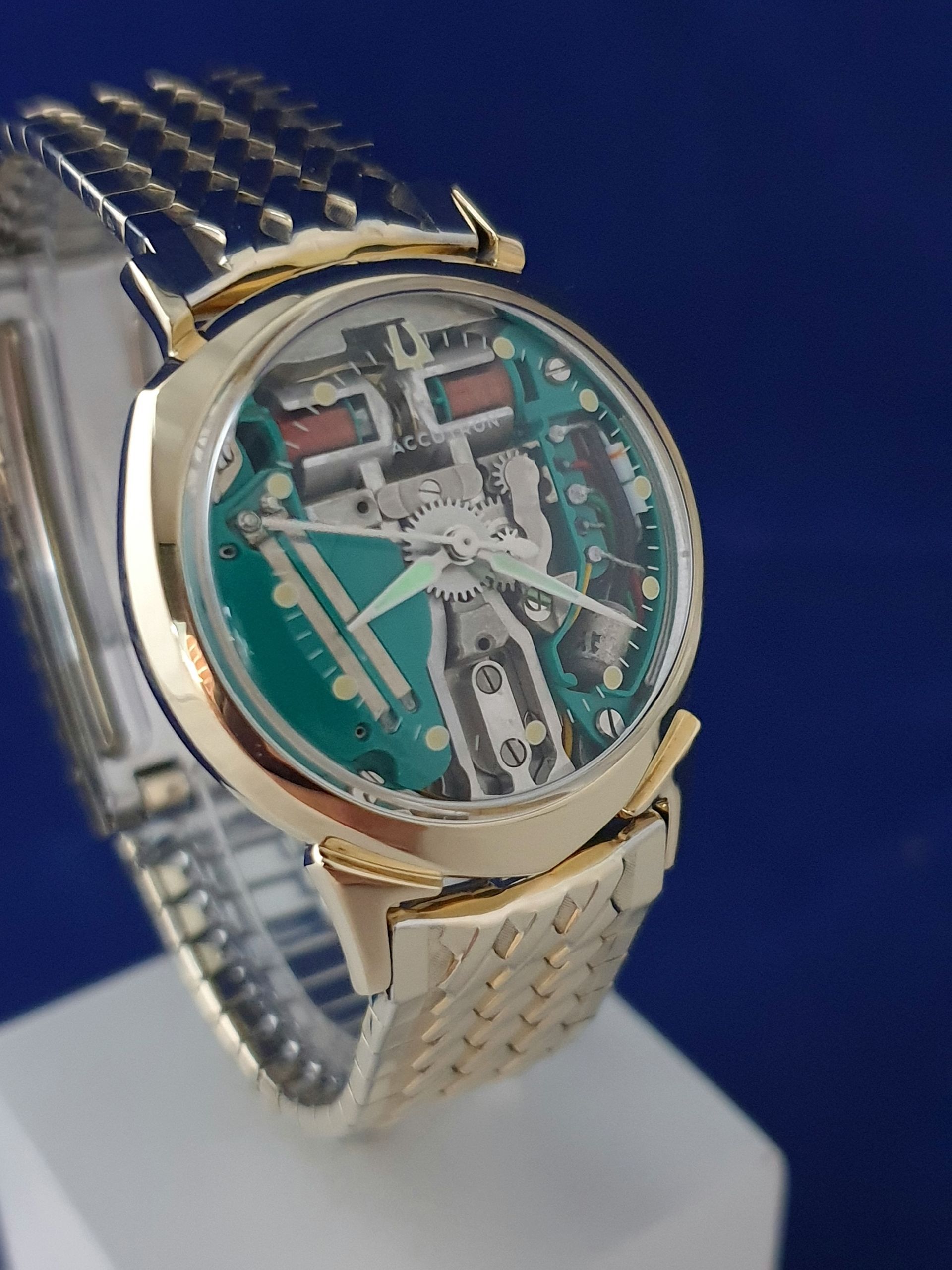 Gold wristwatch with a turquoise skeleton dial and metal band on a white stand against a blue background