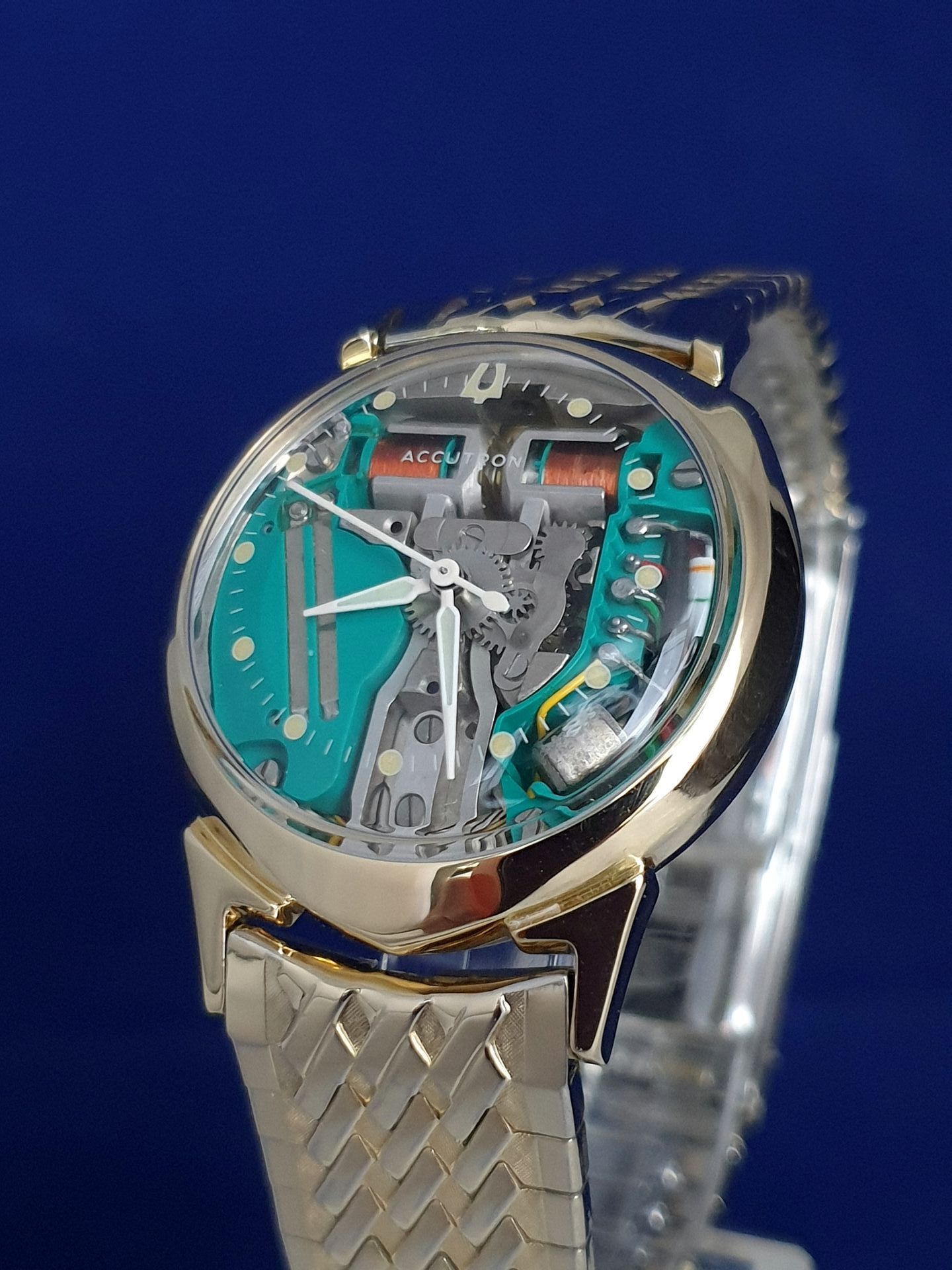 Skeleton wristwatch with turquoise dial and gold mesh band on a blue background